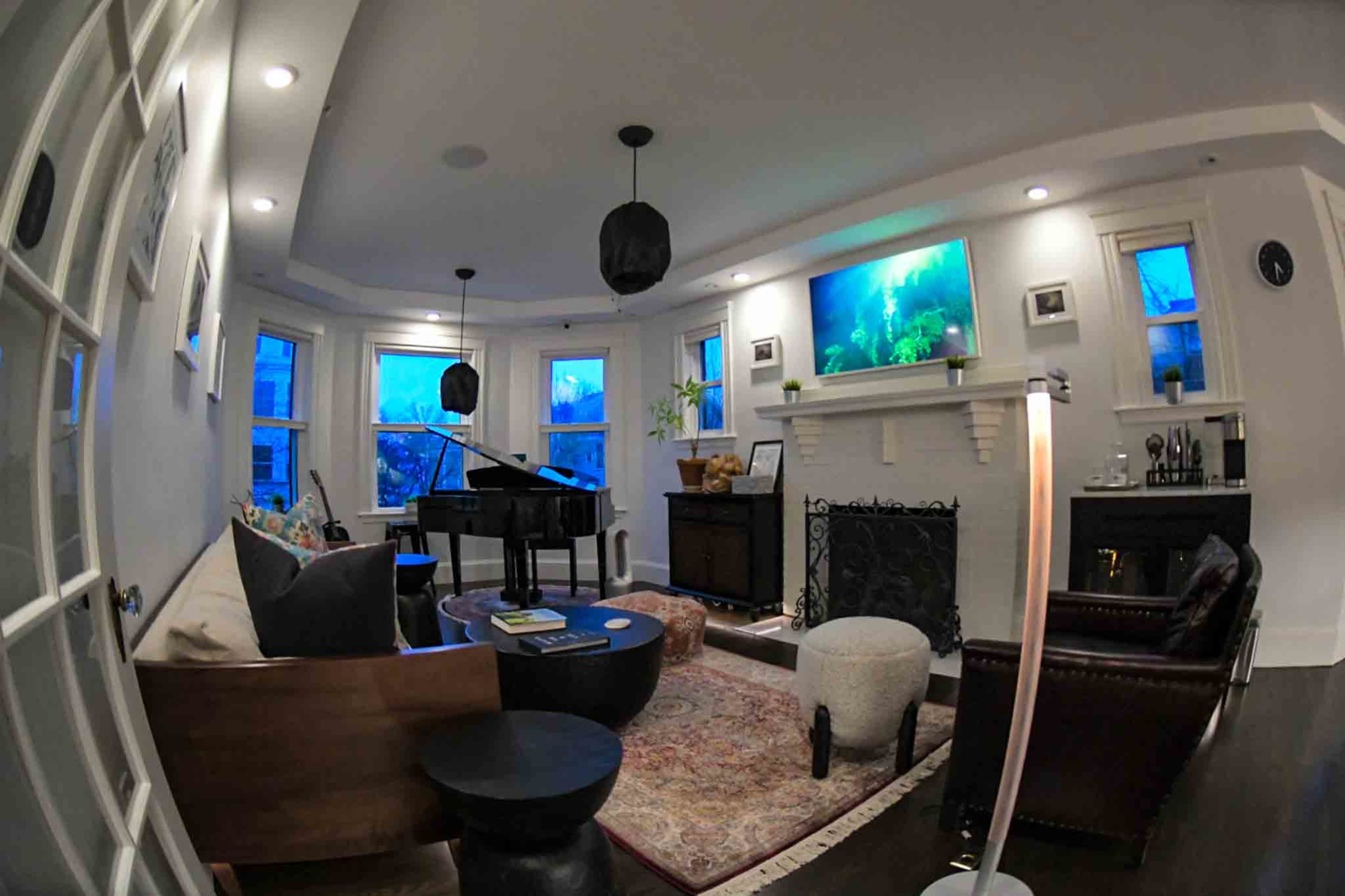 The image shows a cozy living room featuring a piano, a fireplace with a television above it, and large windows allowing natural light to enter.