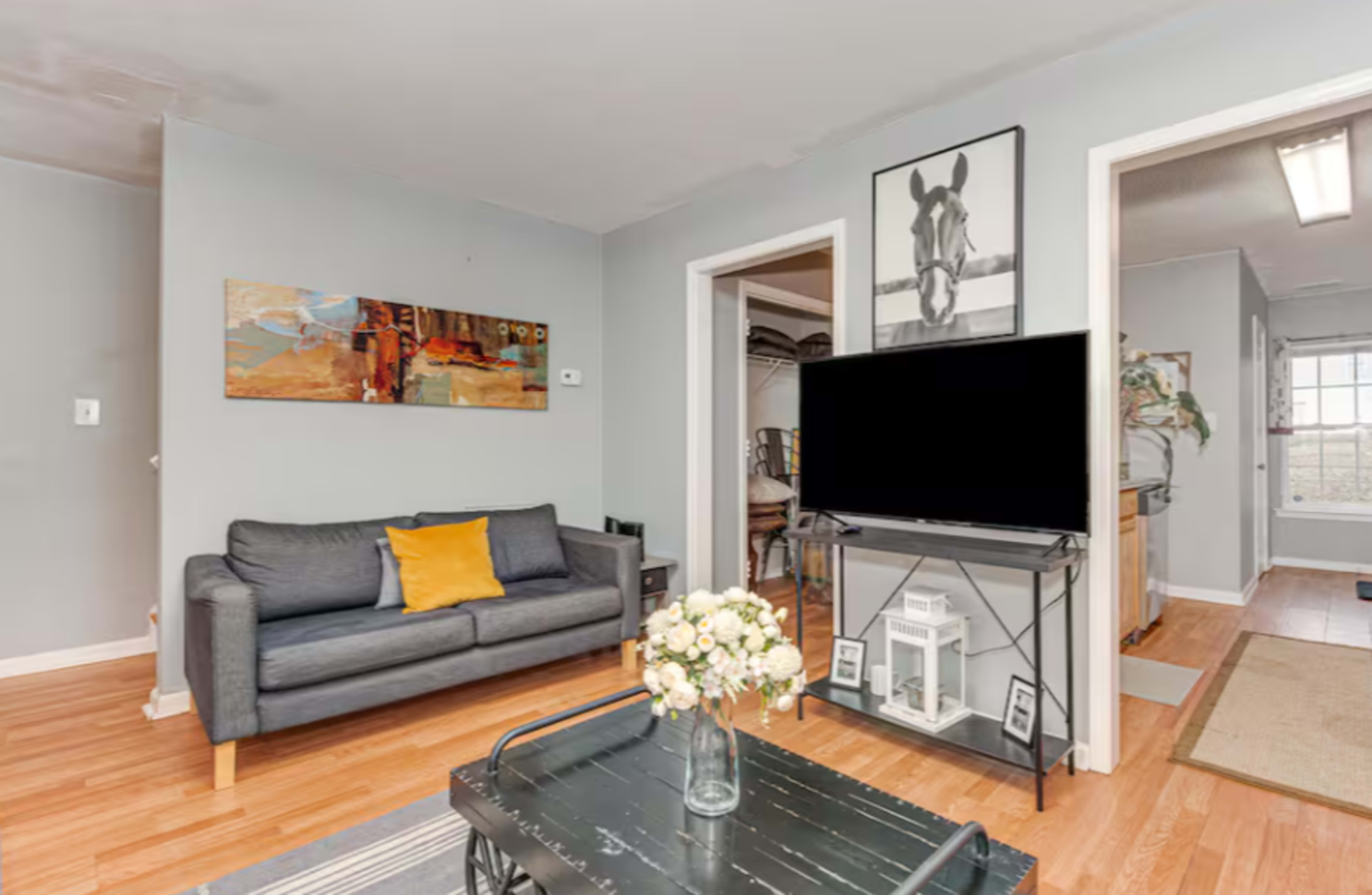 A cozy living room features a gray sofa with a yellow pillow, a black television stand, and a coffee table with a vase of flowers.
