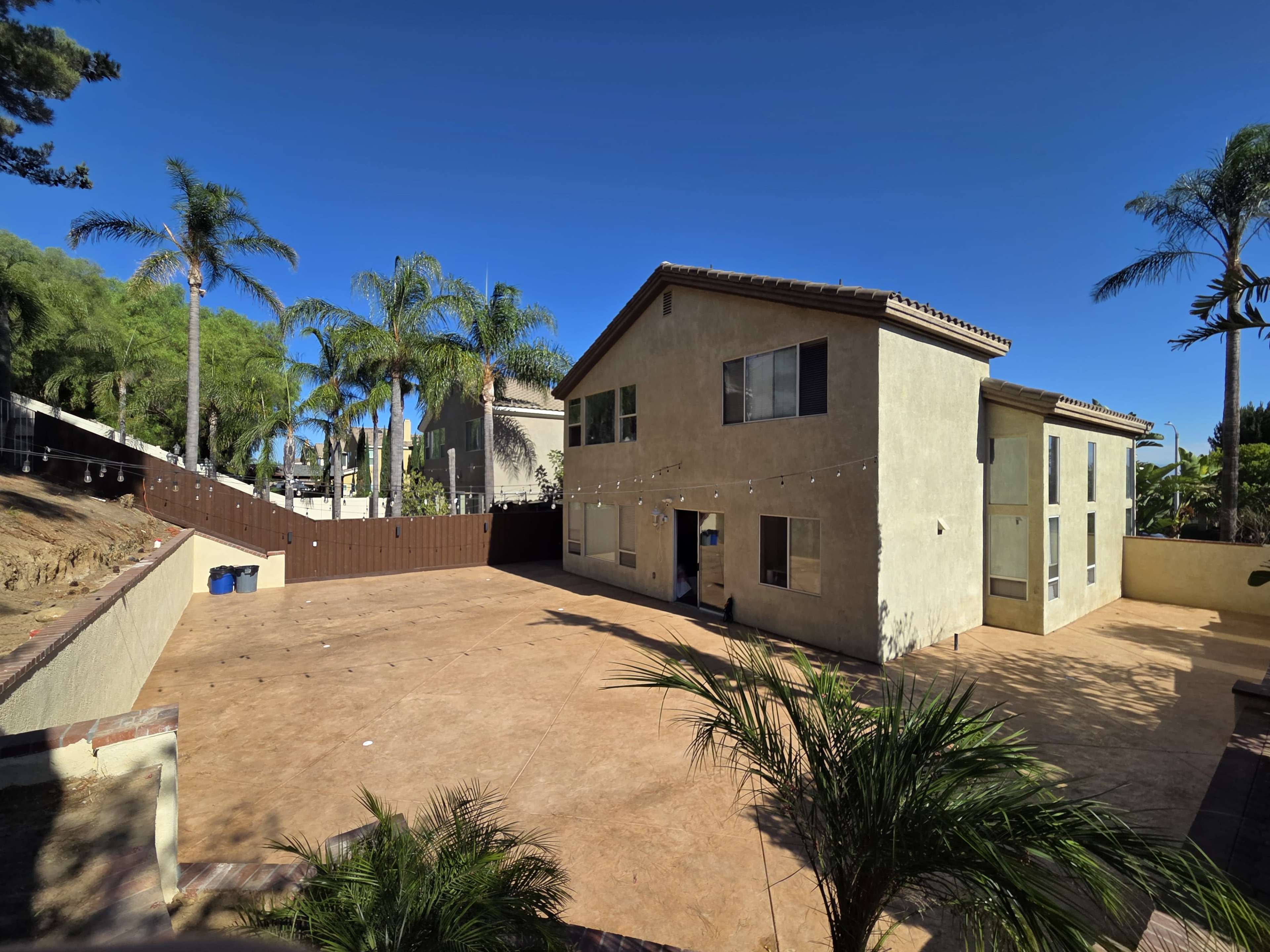 A two-story house with a stucco exterior and a large, empty backyard surrounded by palm trees and a wooden fence is visible in the image.