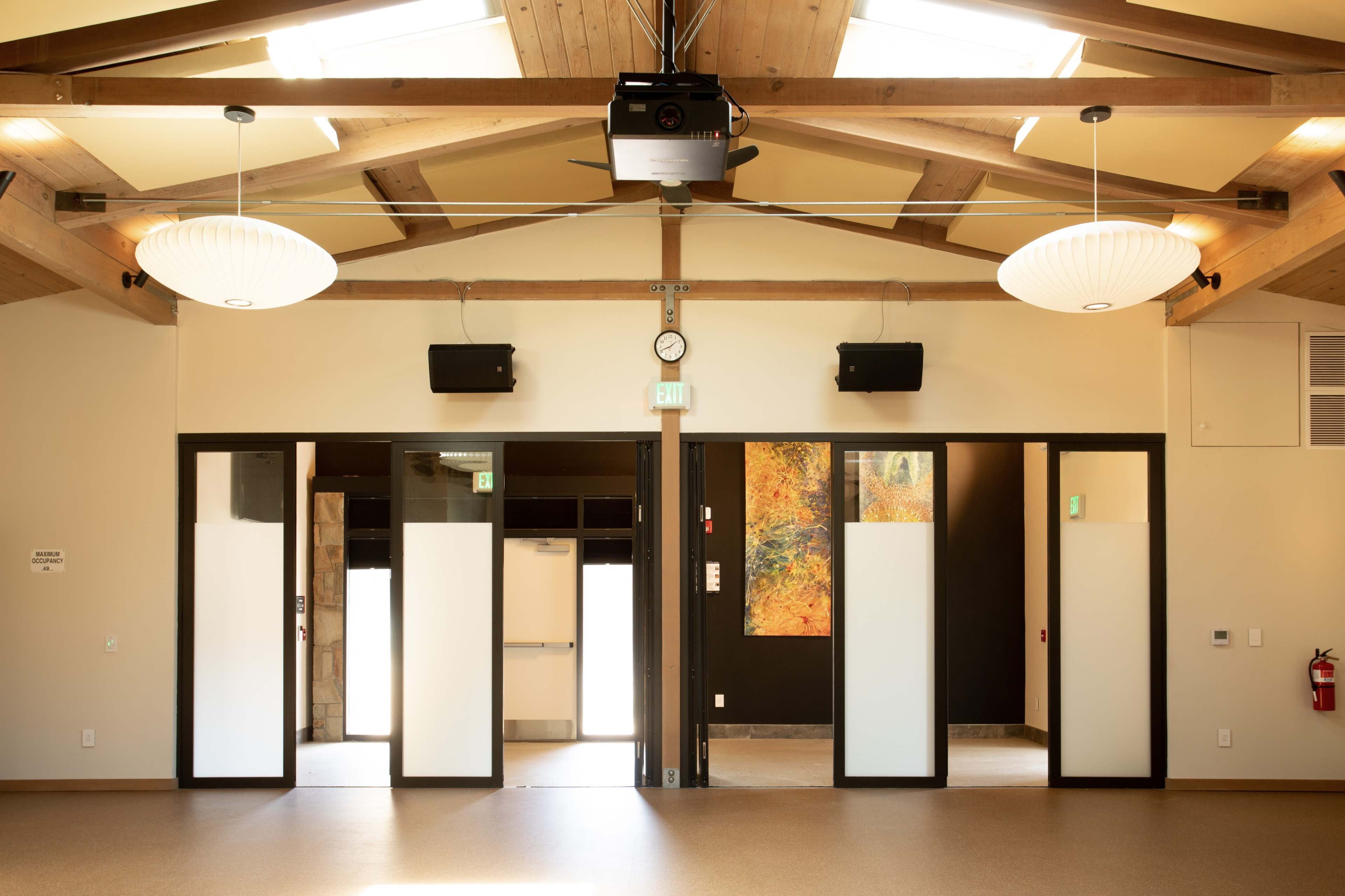 A spacious interior room with wooden beams, large light fixtures, and two sets of double doors leading to adjoining spaces.