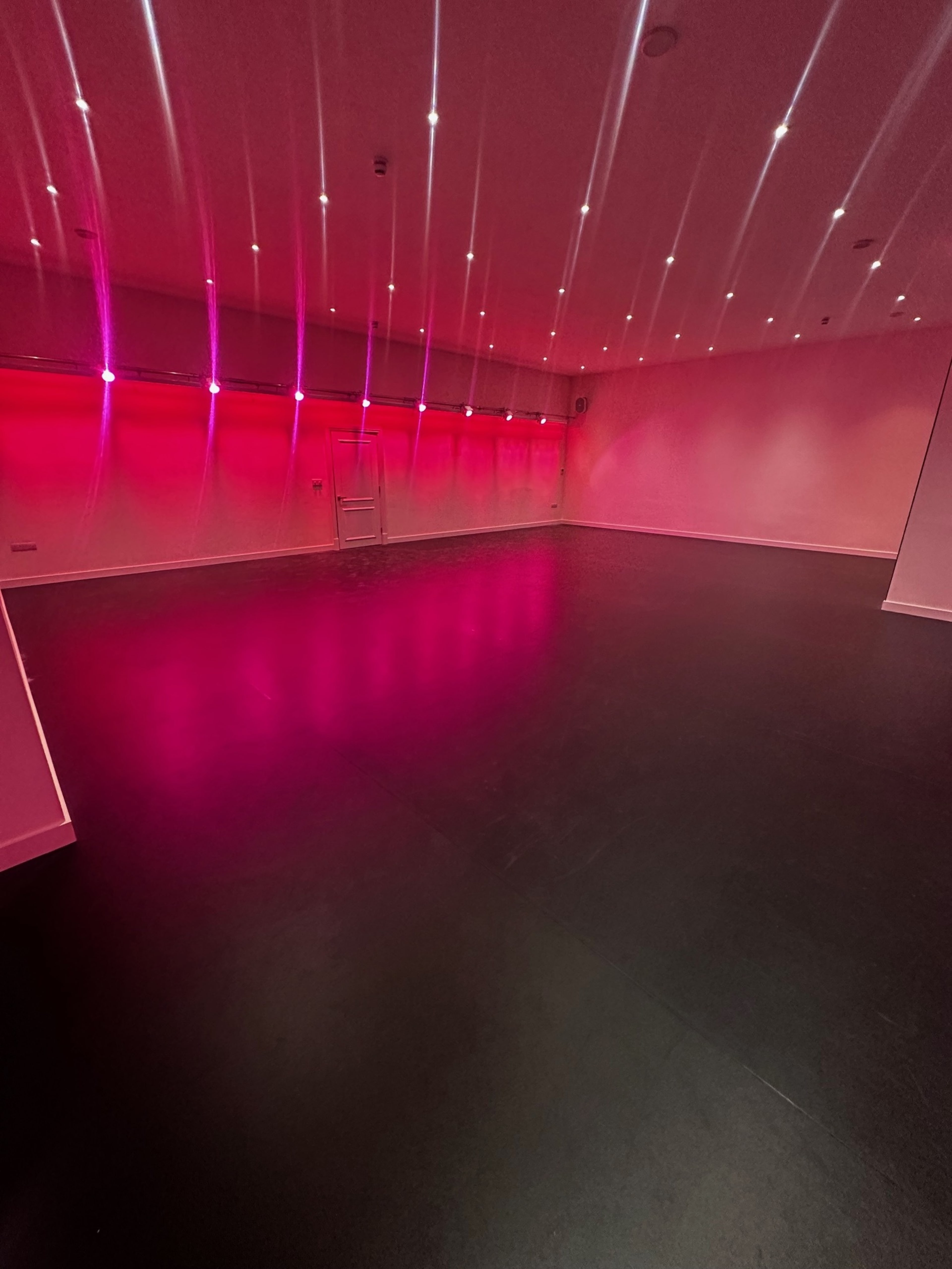 A spacious, empty room features a dark floor and pink lights illuminating the walls.