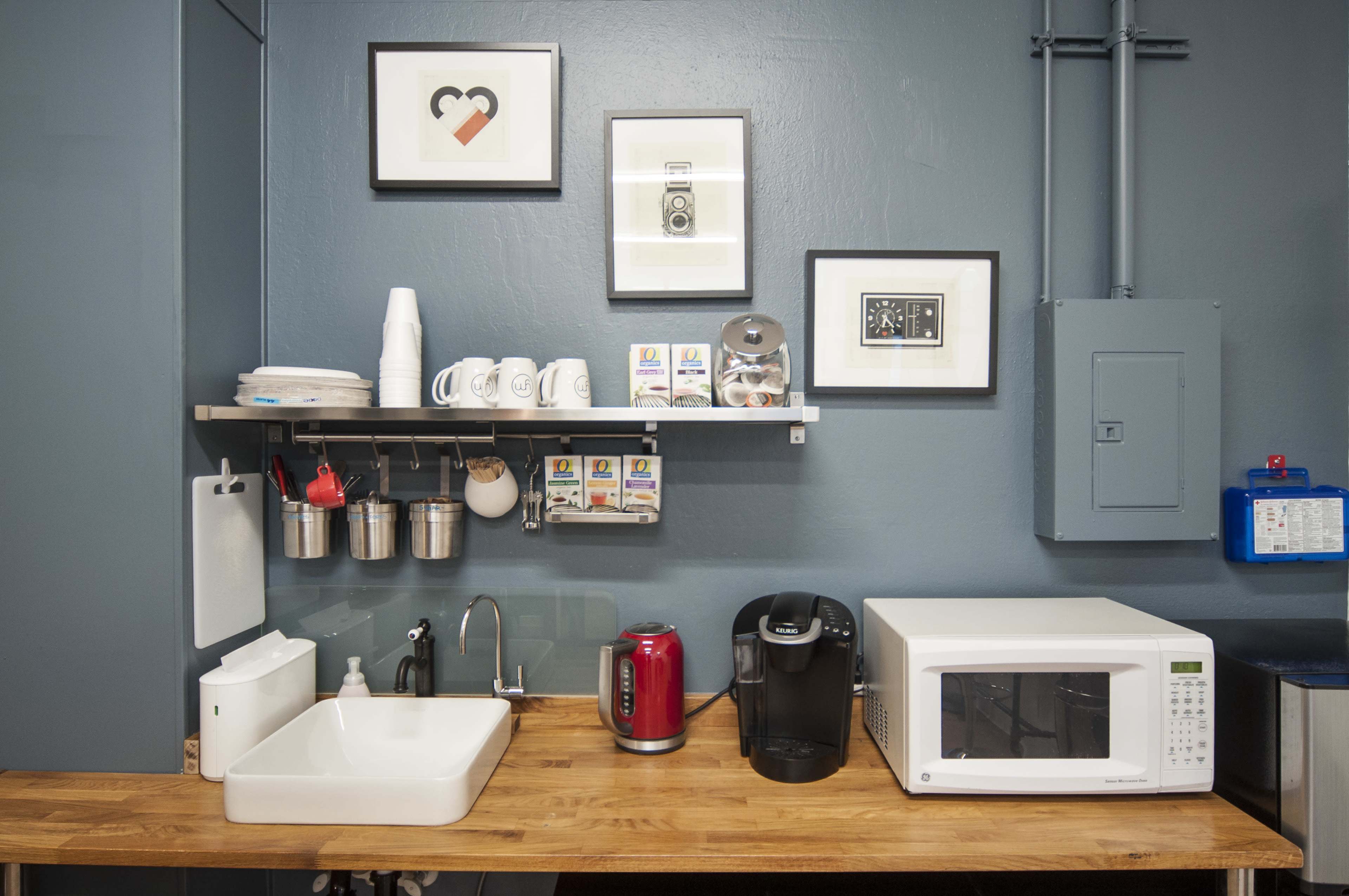 A kitchen area featuring a wooden counter with a sink, a microwave, a coffee maker, and several mugs on a shelf, against a blue wall adorned with framed artwork.