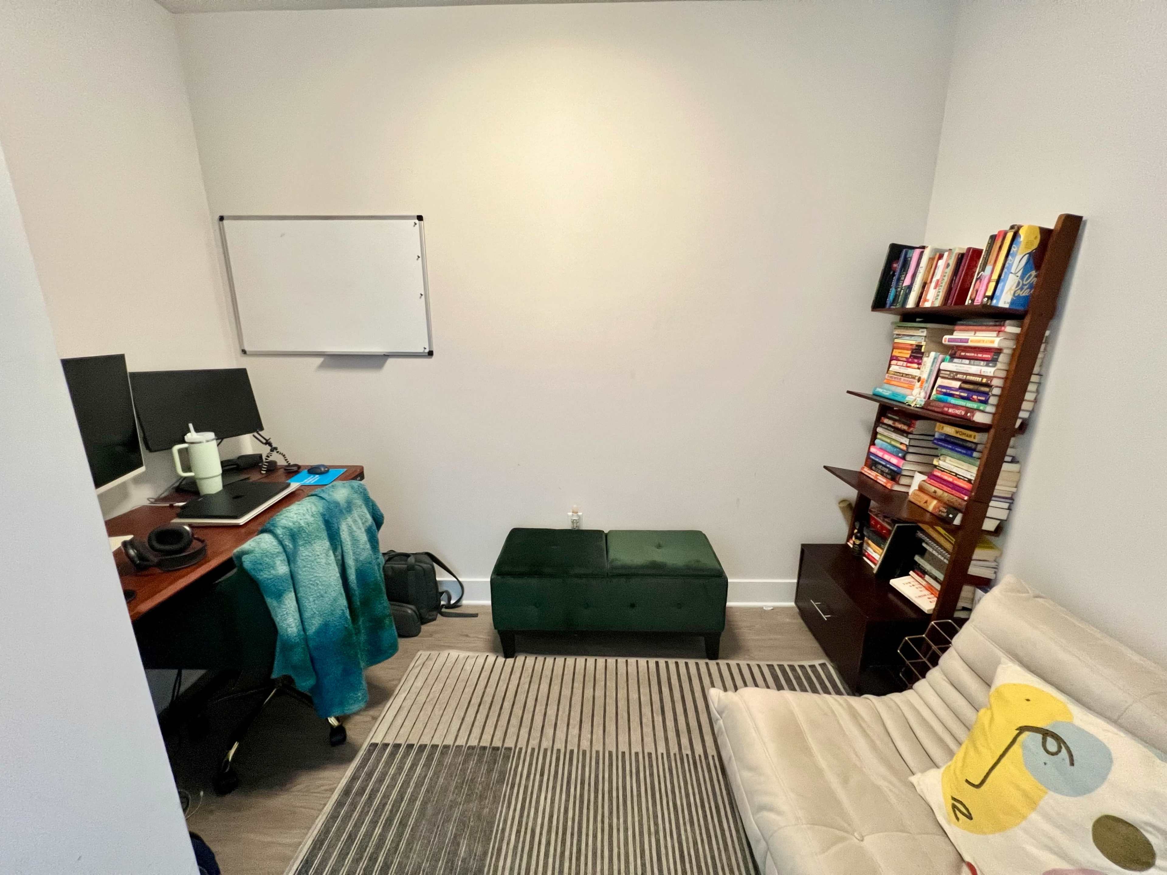 The room features a small workspace with two computer monitors, a green ottoman, a bookshelf filled with books, and a whiteboard on the wall.