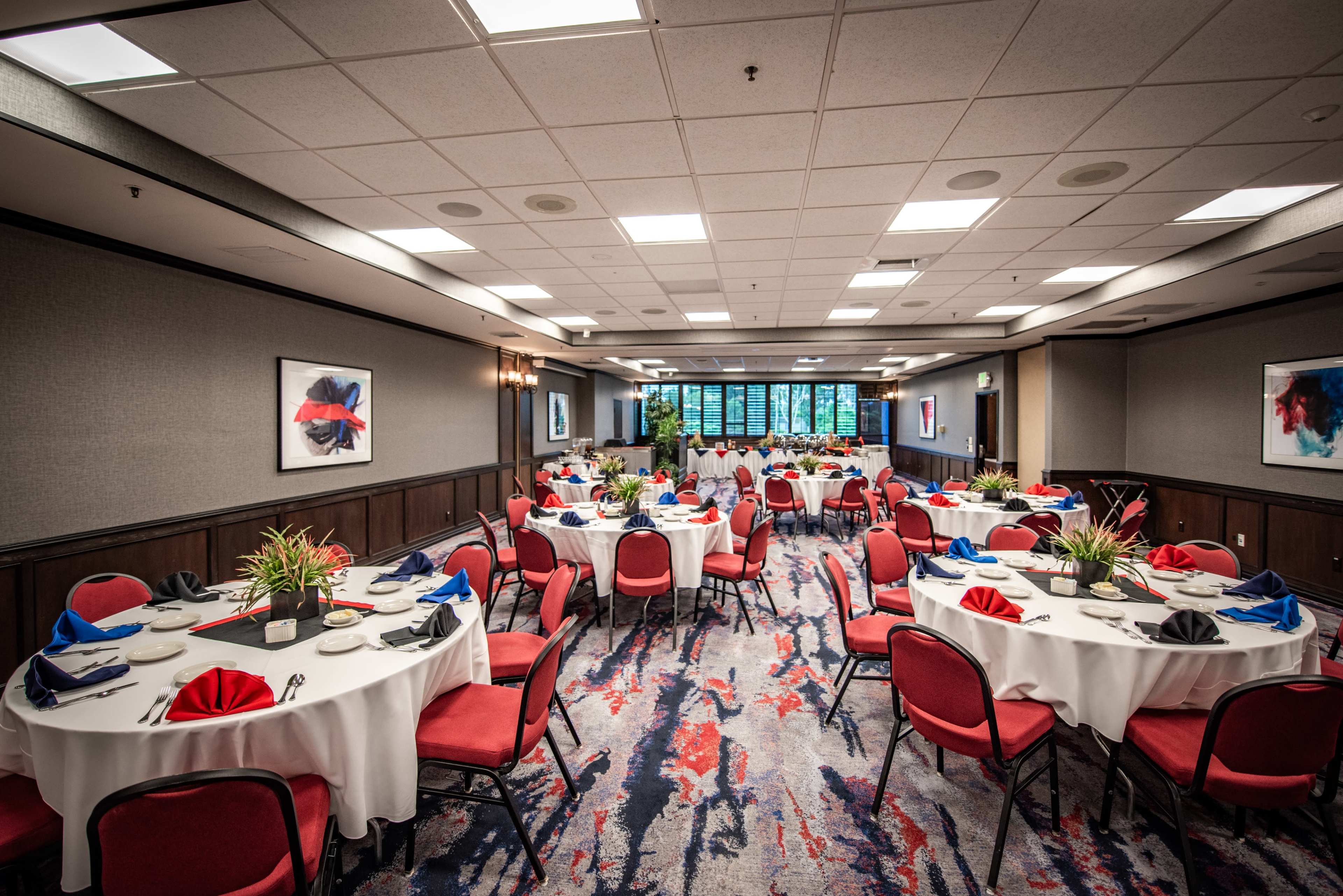 The image shows a banquet room set with round tables covered in white tablecloths, each adorned with red and blue napkins and decorative centerpieces, arranged for an event.