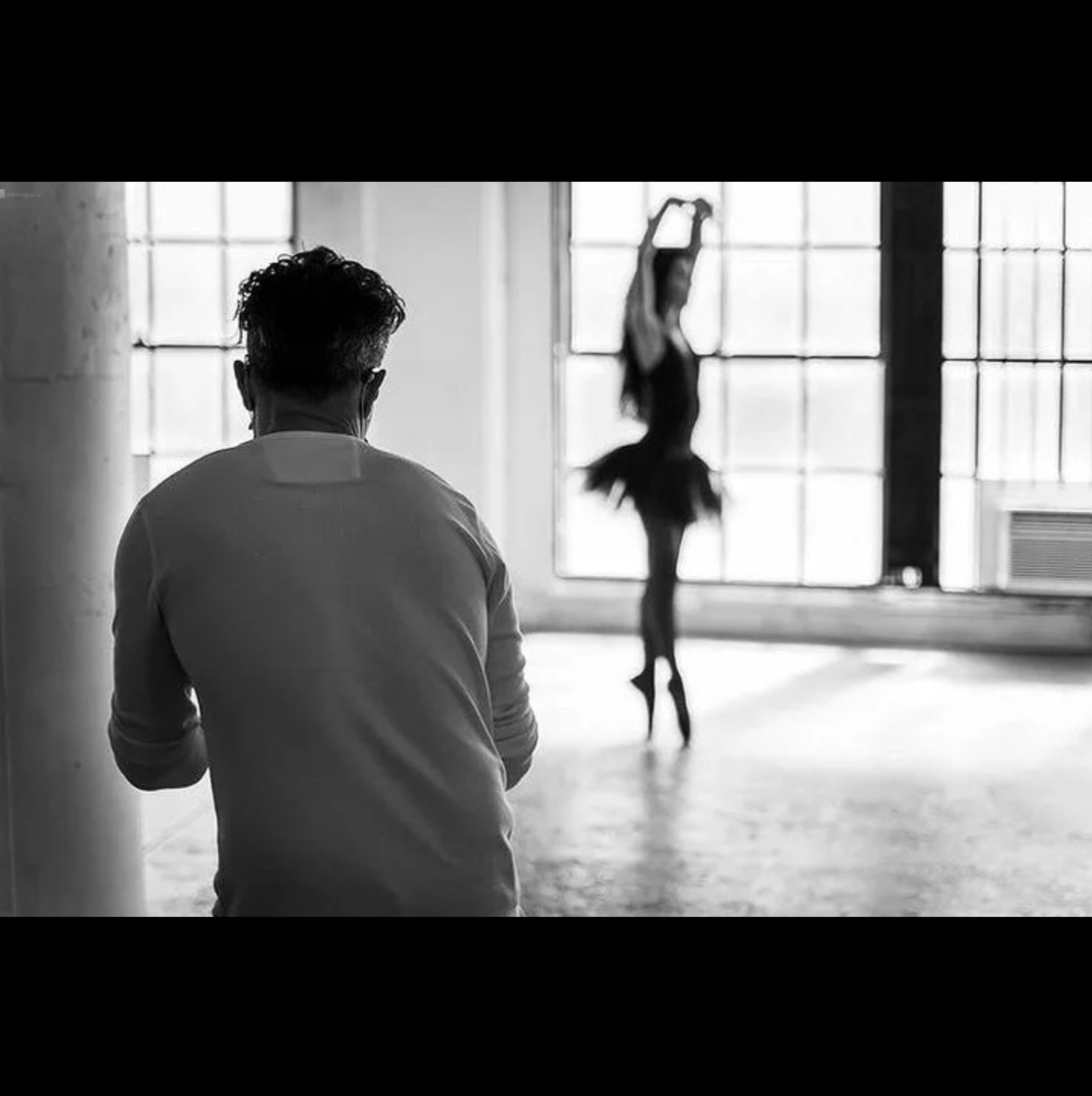 A photographer photographs a ballerina posing in front of large windows in a studio.