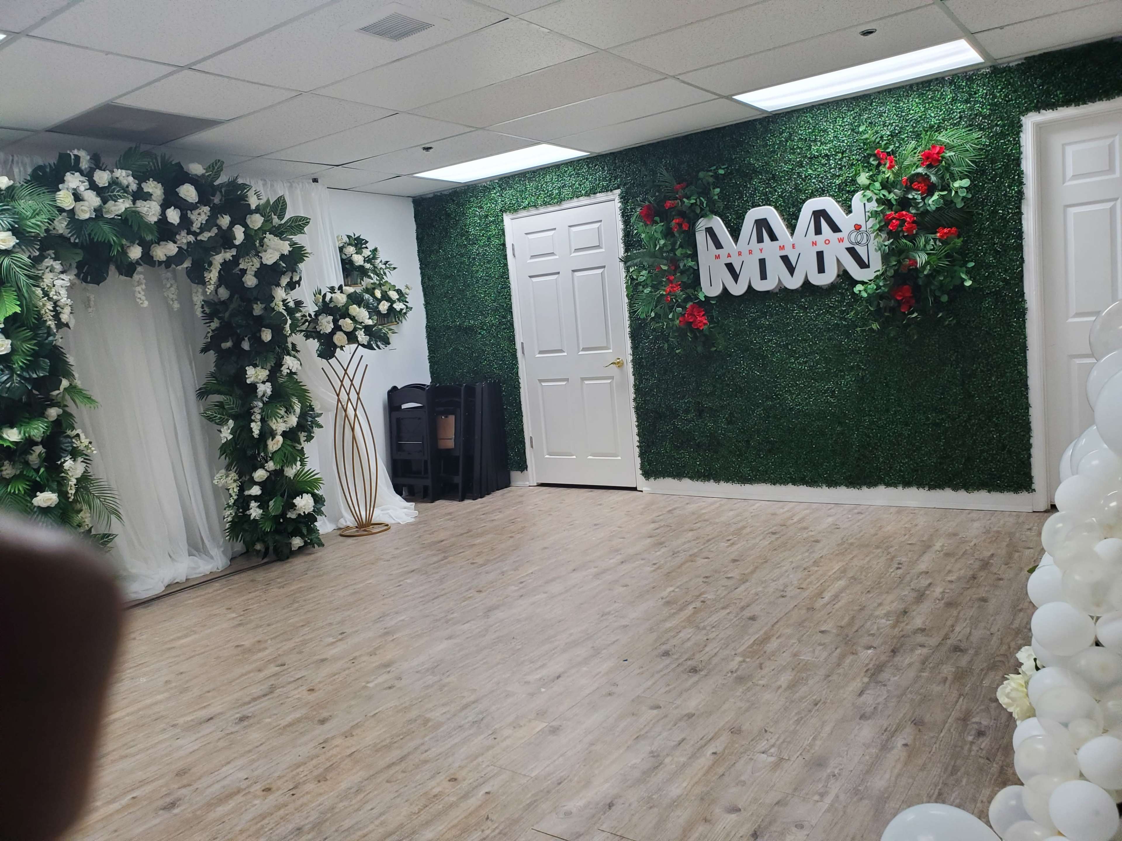The image shows a decorated room with artificial greenery, floral arrangements, a white backdrop, and a logo on the wall.