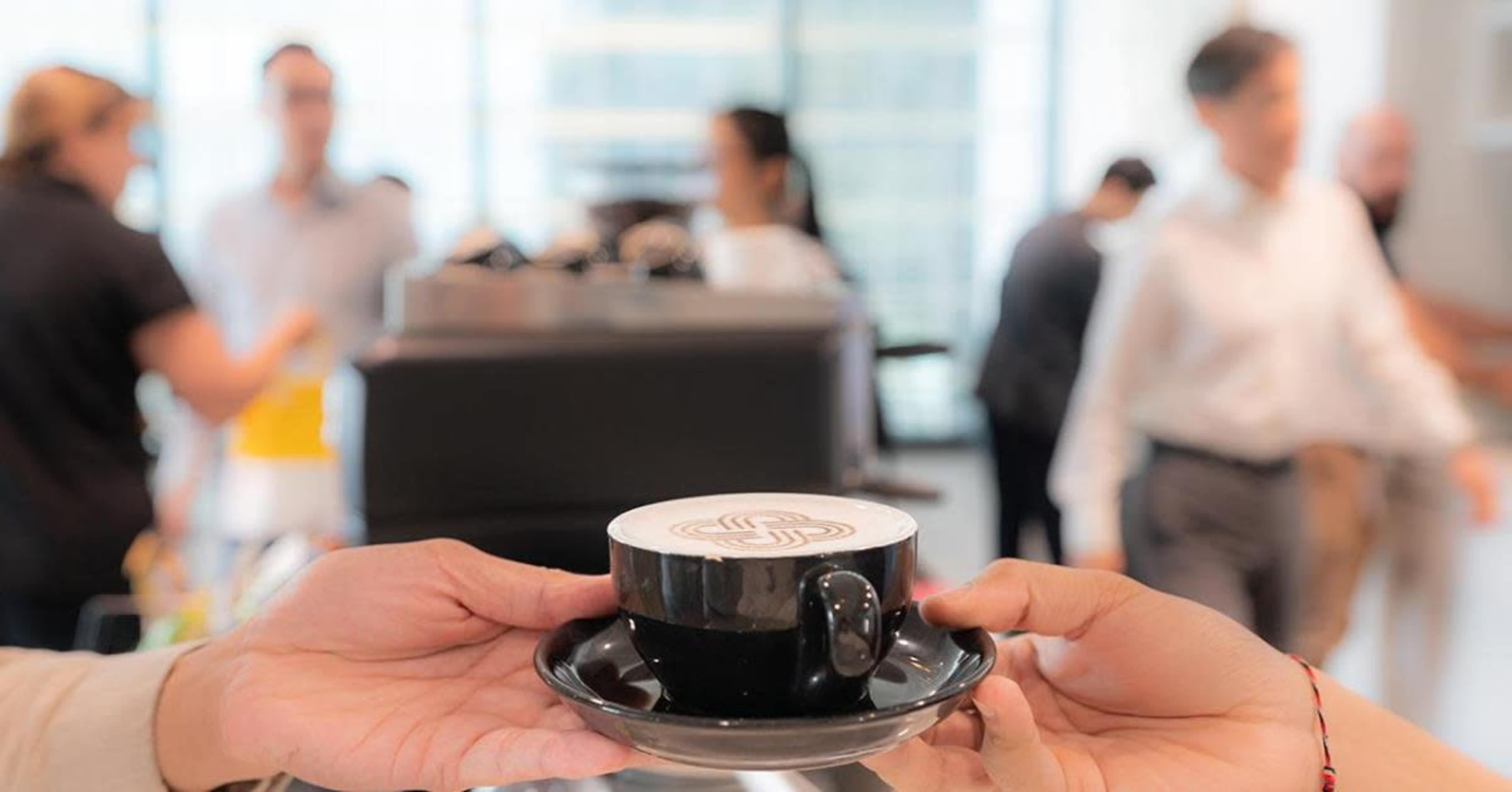 A person hands a black coffee cup with a decorative design on top to another person in a busy café setting.