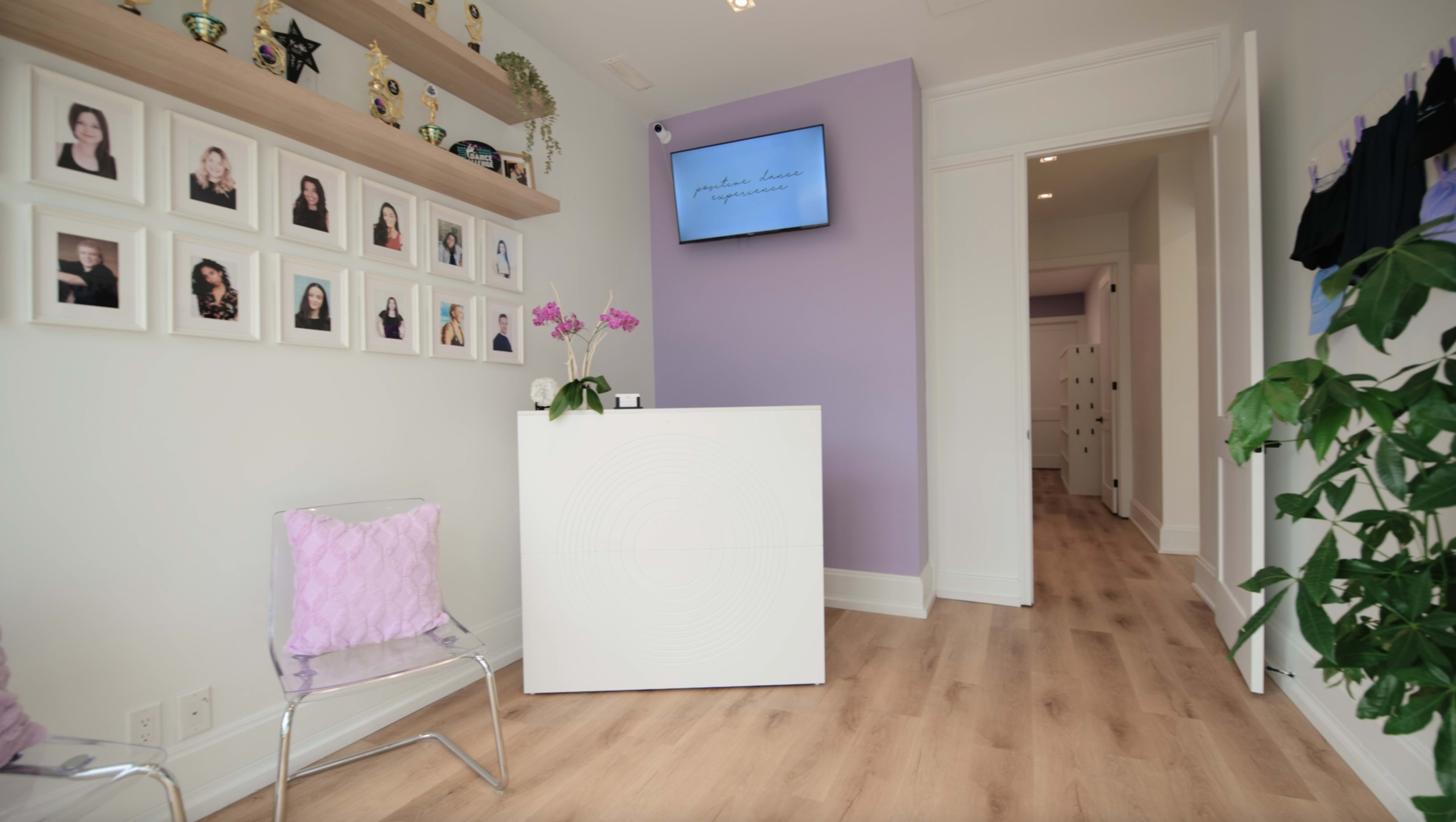 The image shows a welcoming reception area with a white front desk, a lavender accent wall, framed photographs on the shelves, and a display screen, with a hallway leading to another room.