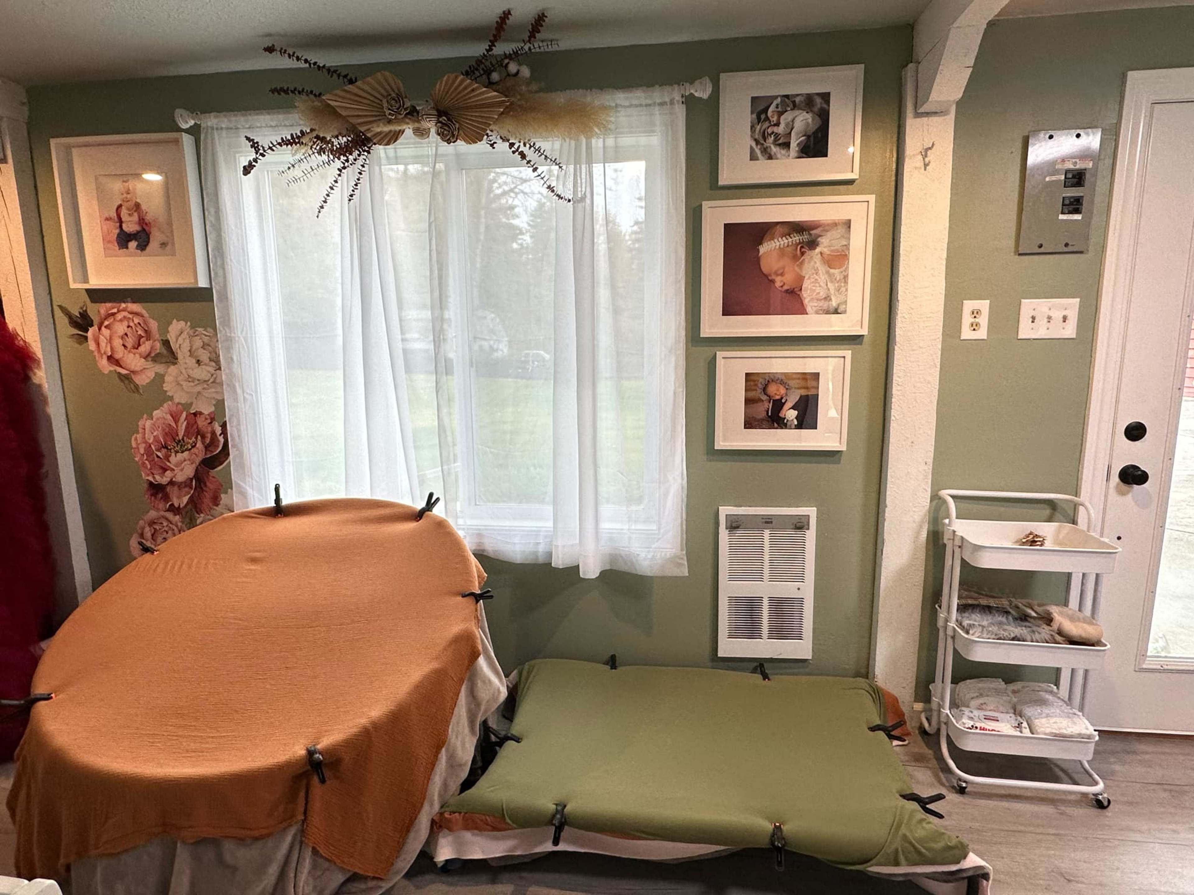 A room features two makeshift beds covered with orange and green bedding, a window adorned with sheer curtains, and several framed photographs on the walls.