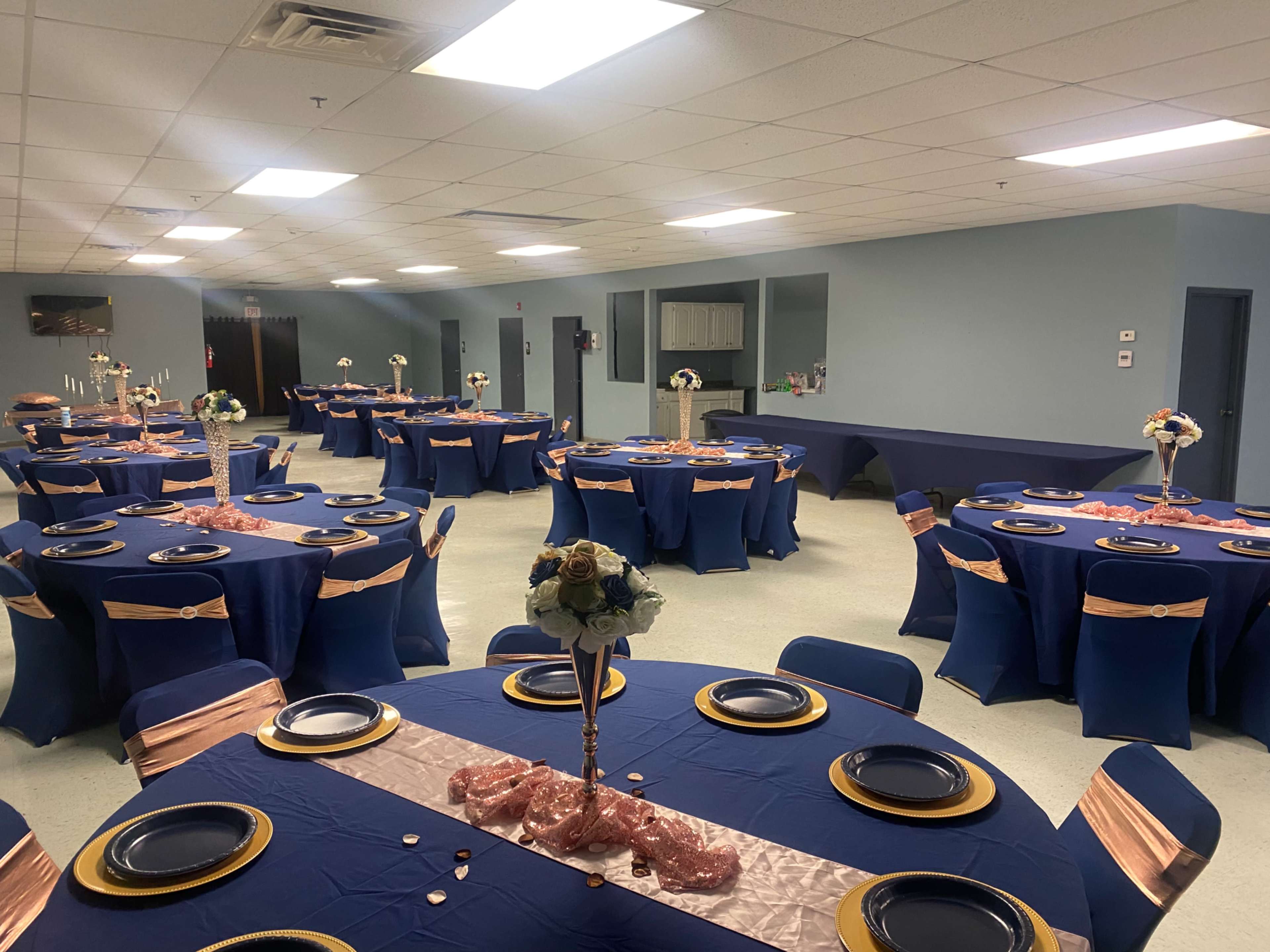 A banquet hall set with several round tables covered in navy blue tablecloths and adorned with pink sashes, each featuring gold plates and floral centerpieces.