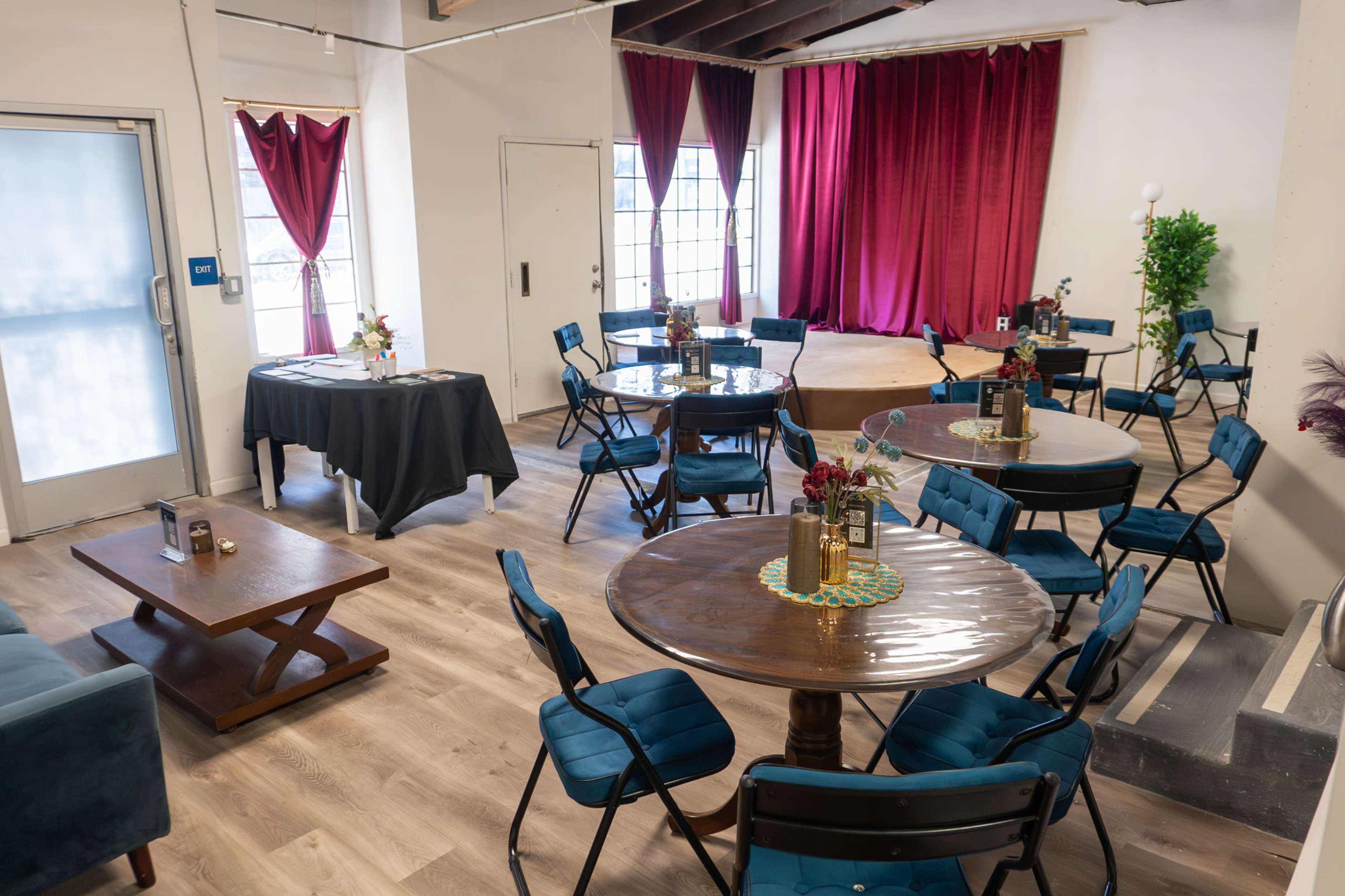 The image shows a room with several round tables and chairs arranged around a wooden floor, featuring red curtains and a small stage area at the front.