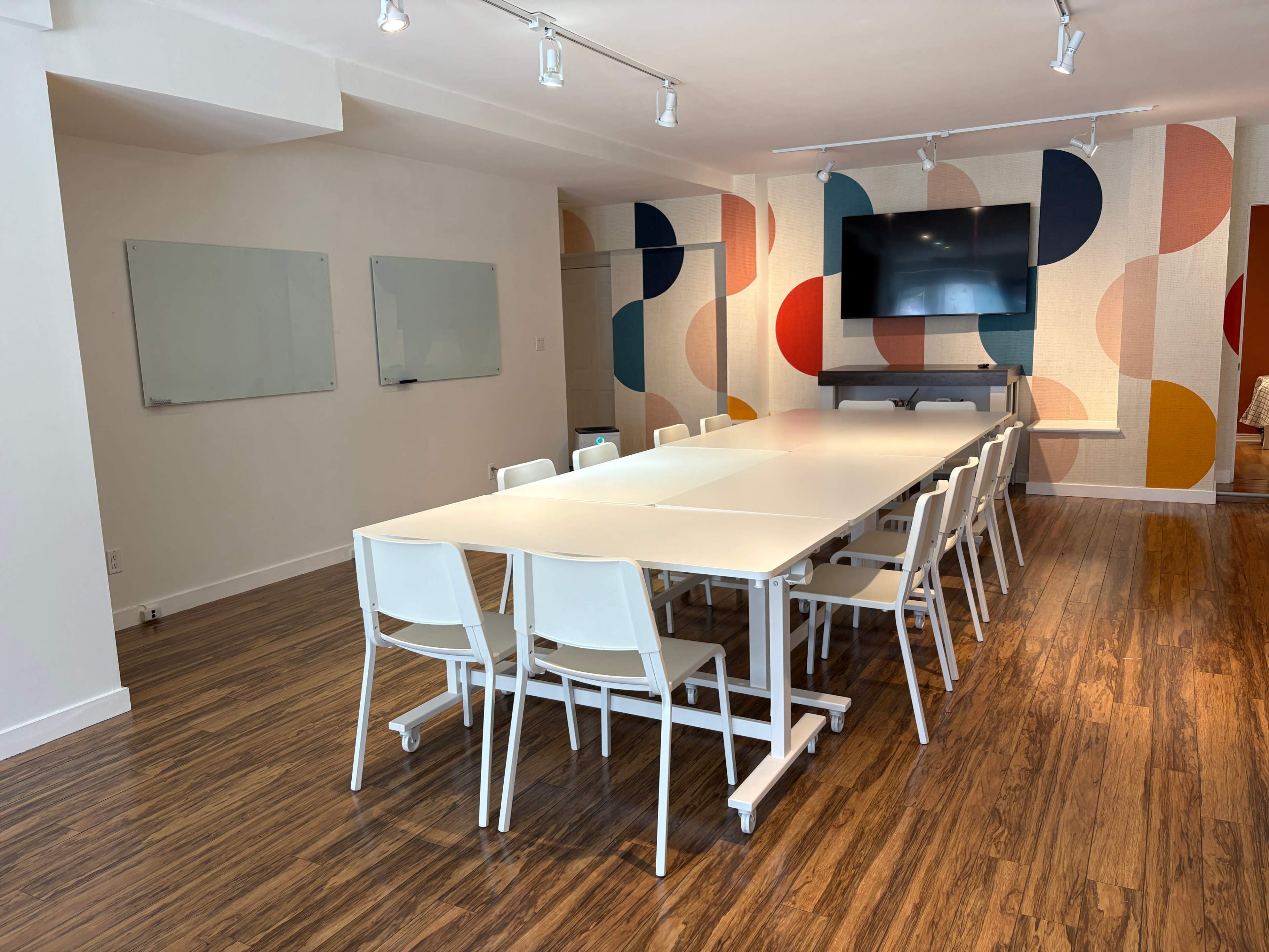 The image shows a modern conference room with a long white table surrounded by white chairs, large whiteboards on the walls, and a colorful accent wall featuring geometric patterns.
