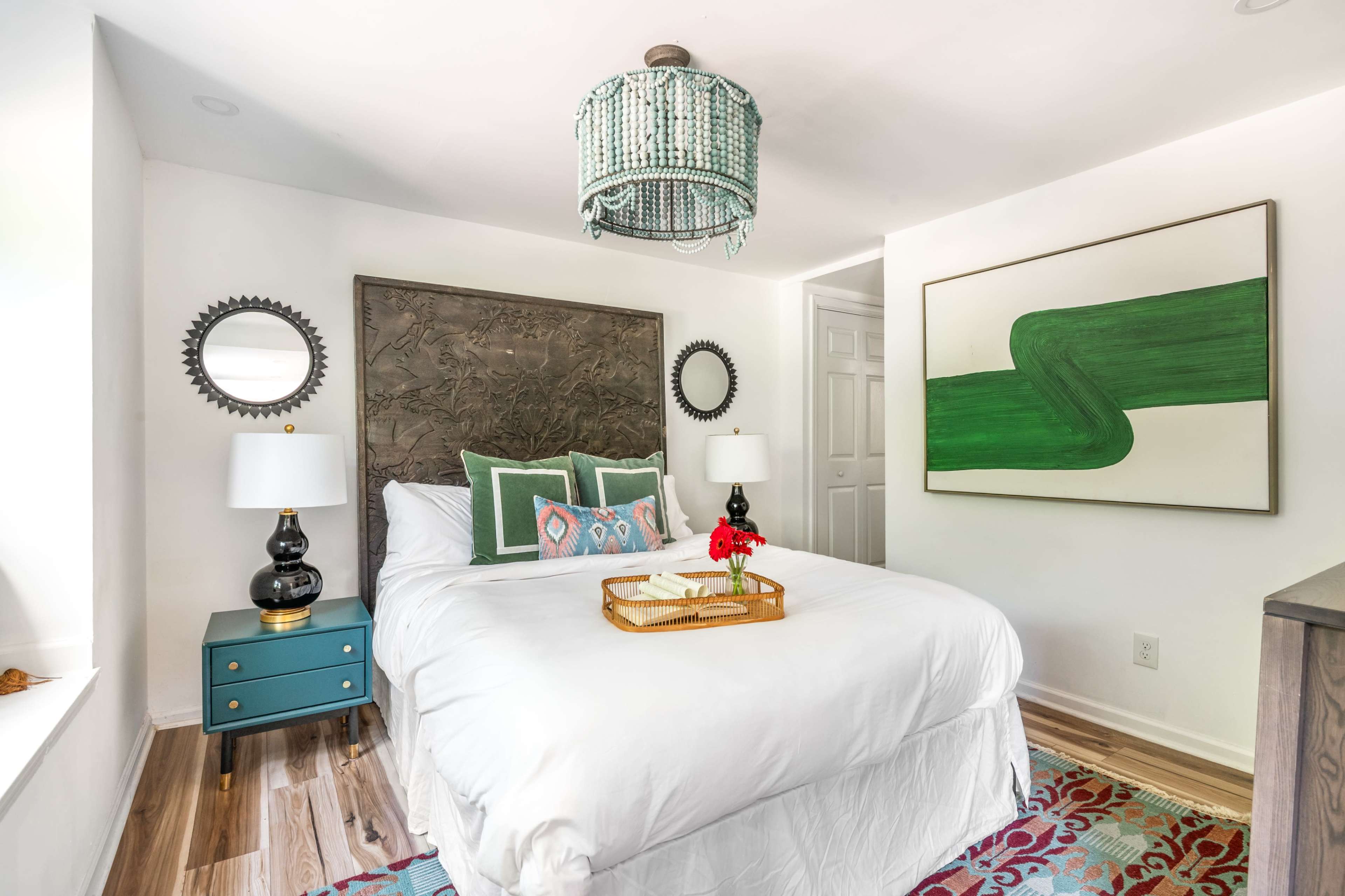 The image shows a neatly arranged bedroom featuring a bed with white bedding, a decorative wall art piece, and a turquoise nightstand with lamps on either side.