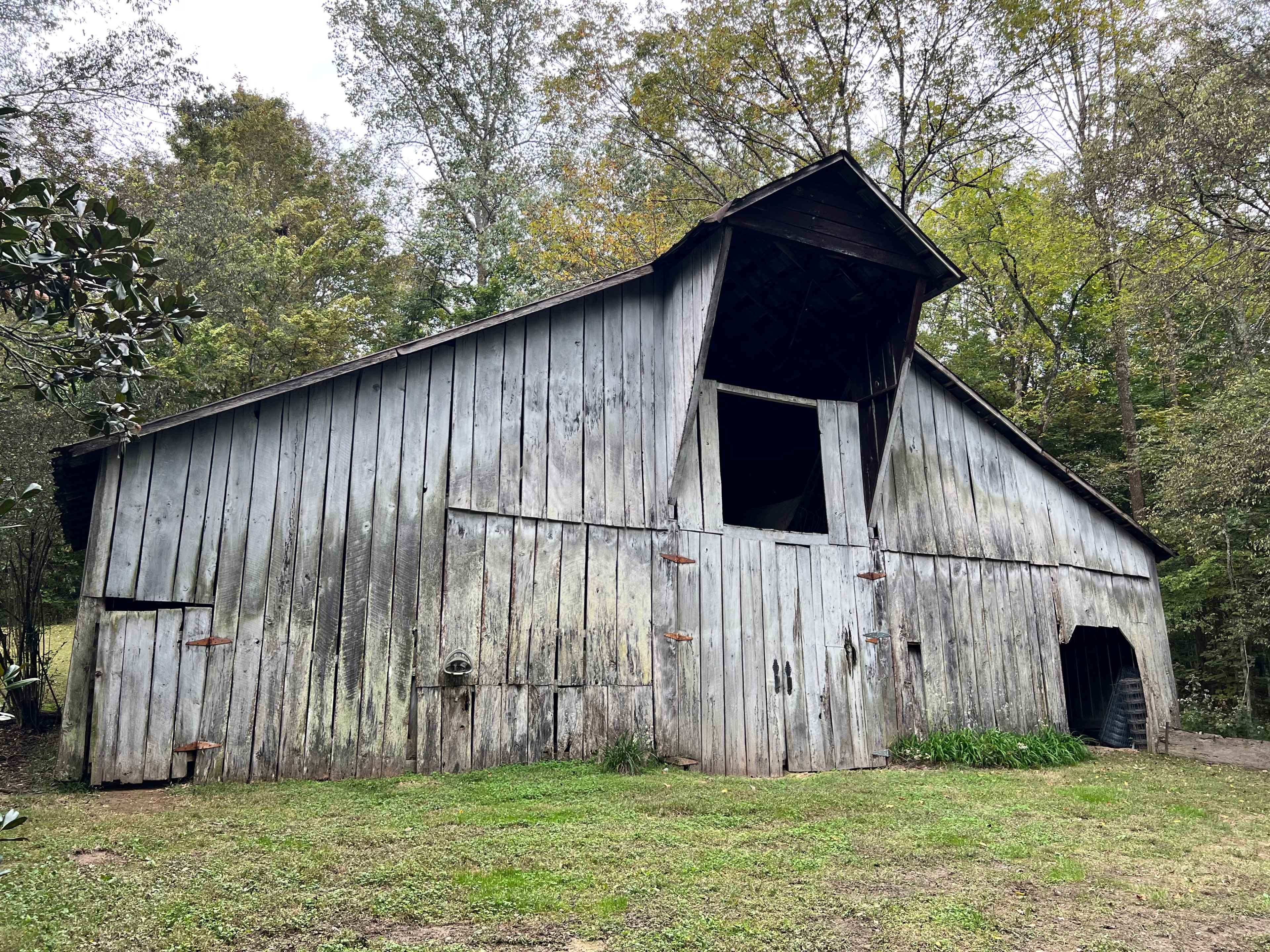 A weathered wooden barn stands at an angle in a wooded area, with green grass and trees surrounding it.