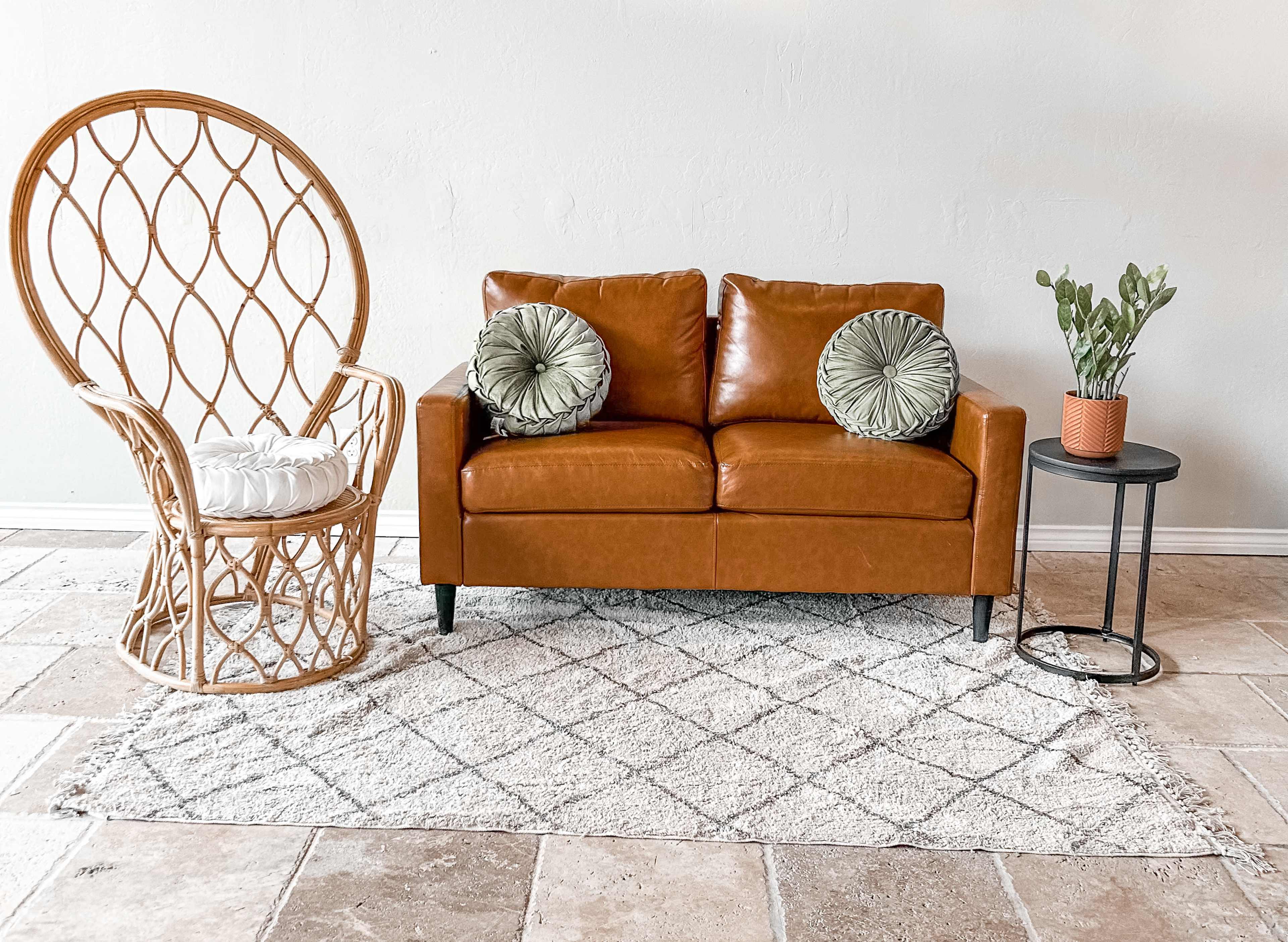 A brown leather sofa with two cushions, a rattan chair, a small side table, and a potted plant, all arranged on a patterned rug in a neutral-colored room.