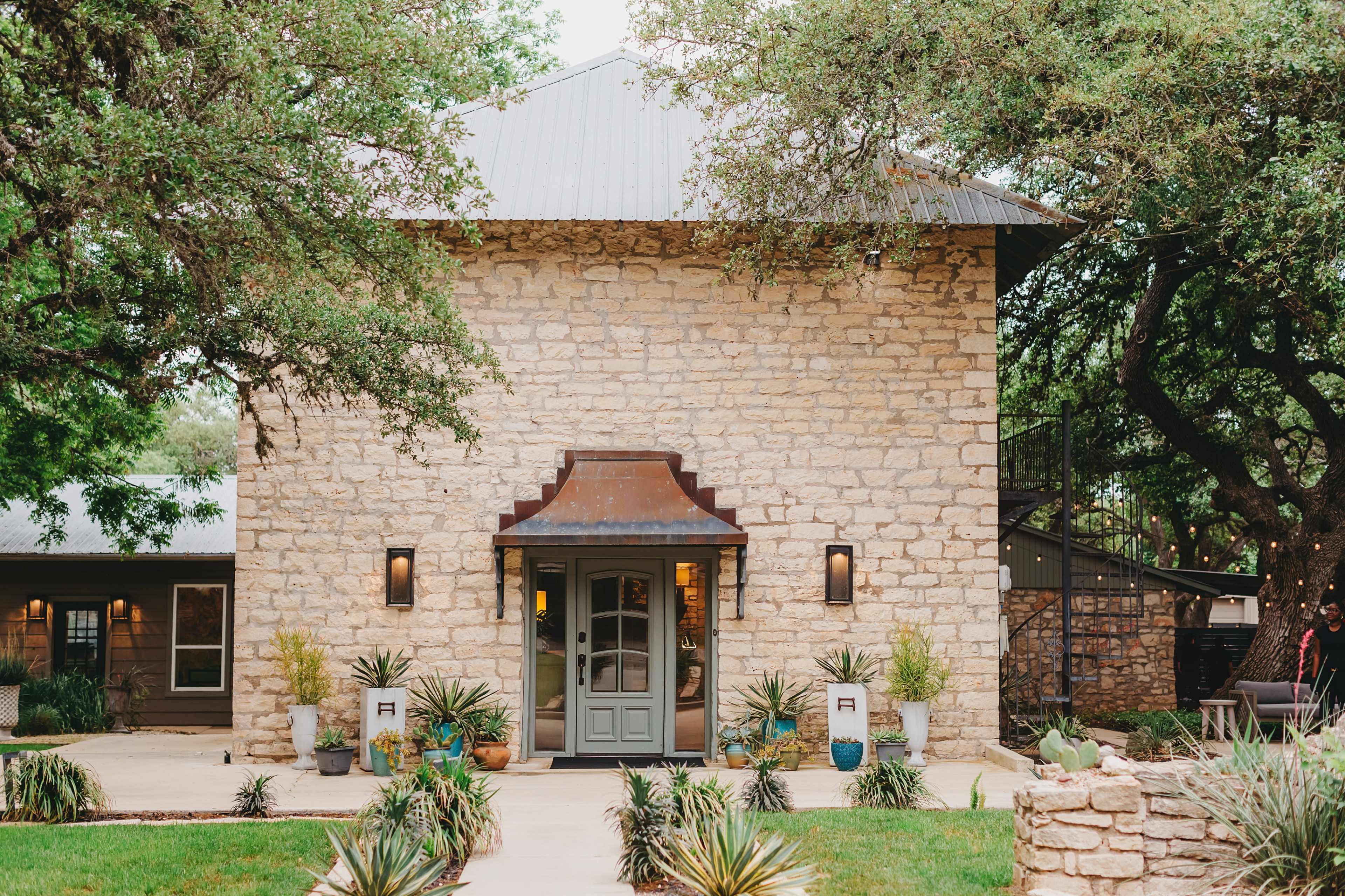 The image shows a stone building with a metal roof, framed by trees and landscaped with potted plants.