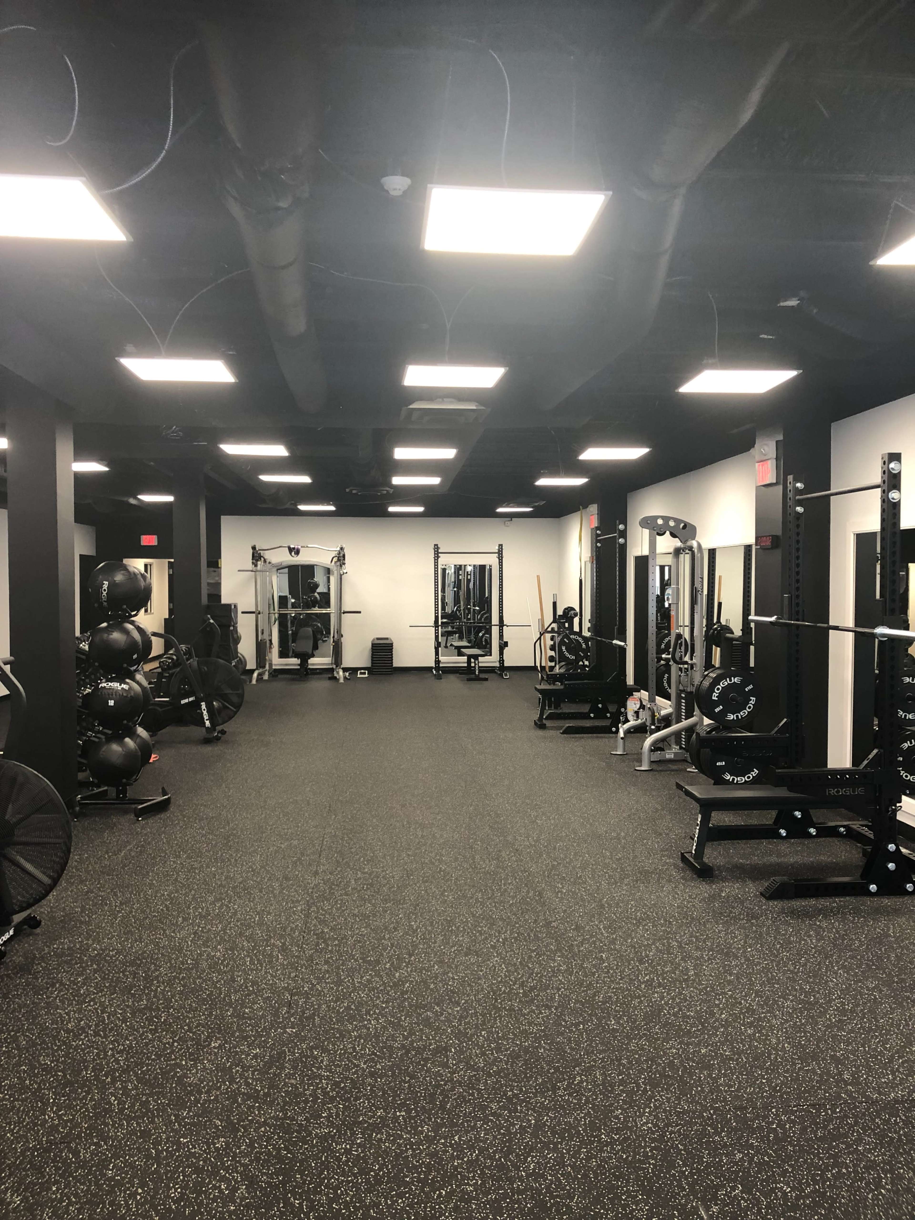 The image shows a spacious gym interior featuring various weightlifting equipment, exercise machines, and workout areas on a rubberized floor.