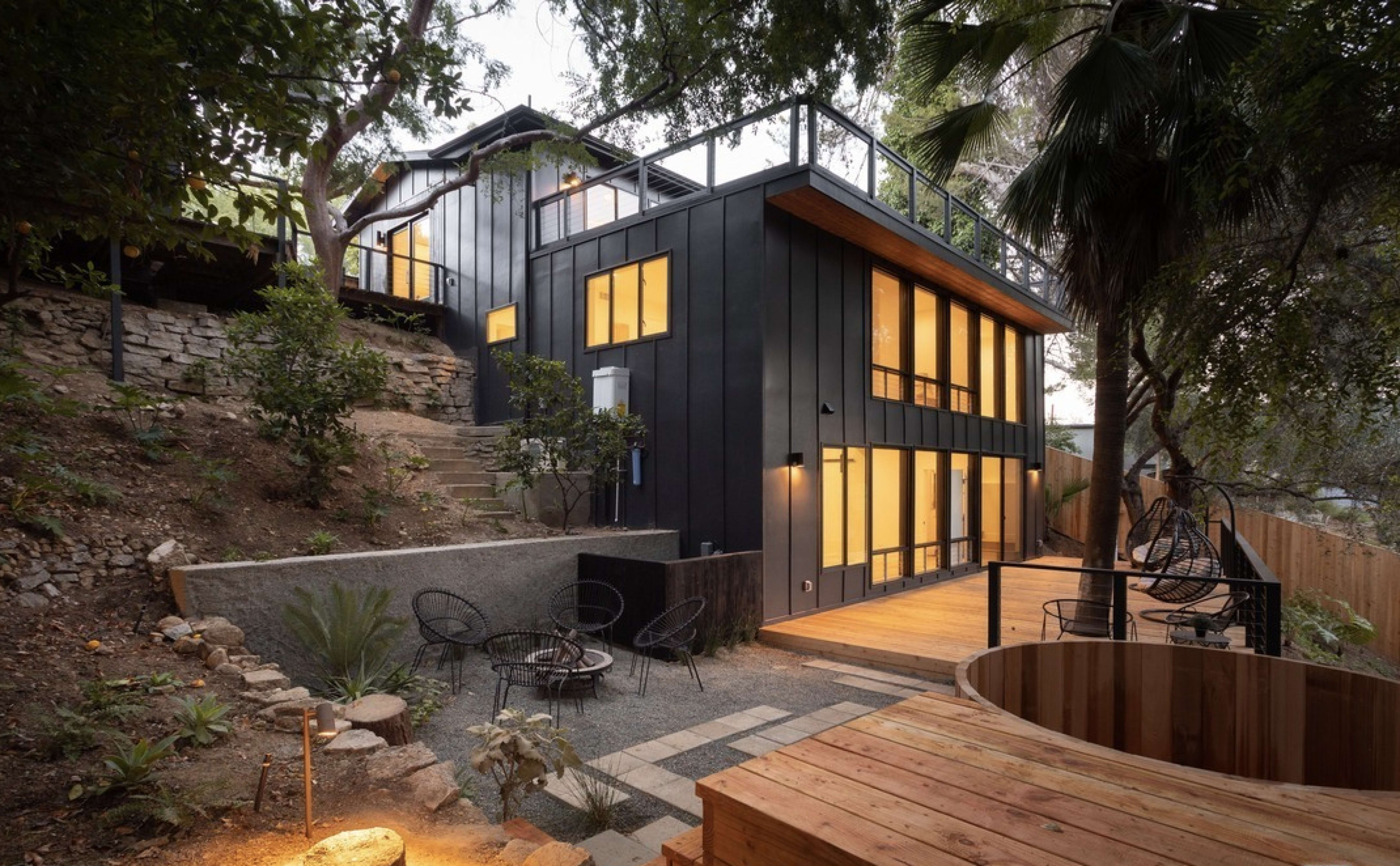A modern hillside house with large windows, a wooden deck, and a hot tub, surrounded by trees and landscaped stone pathways.