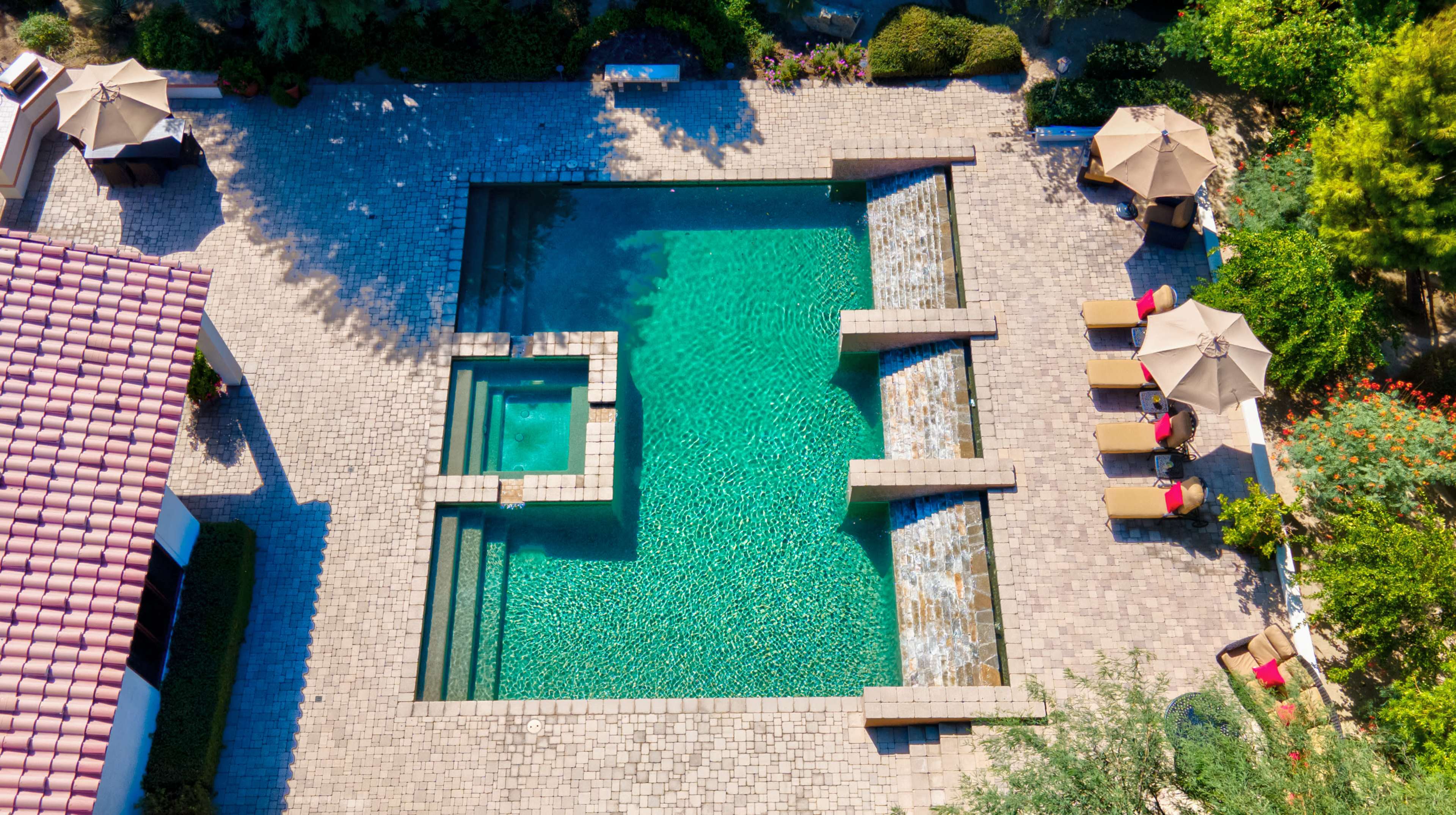 An aerial view of a rectangular swimming pool surrounded by stone decking, with a smaller hot tub and several lounge chairs and umbrellas nearby.