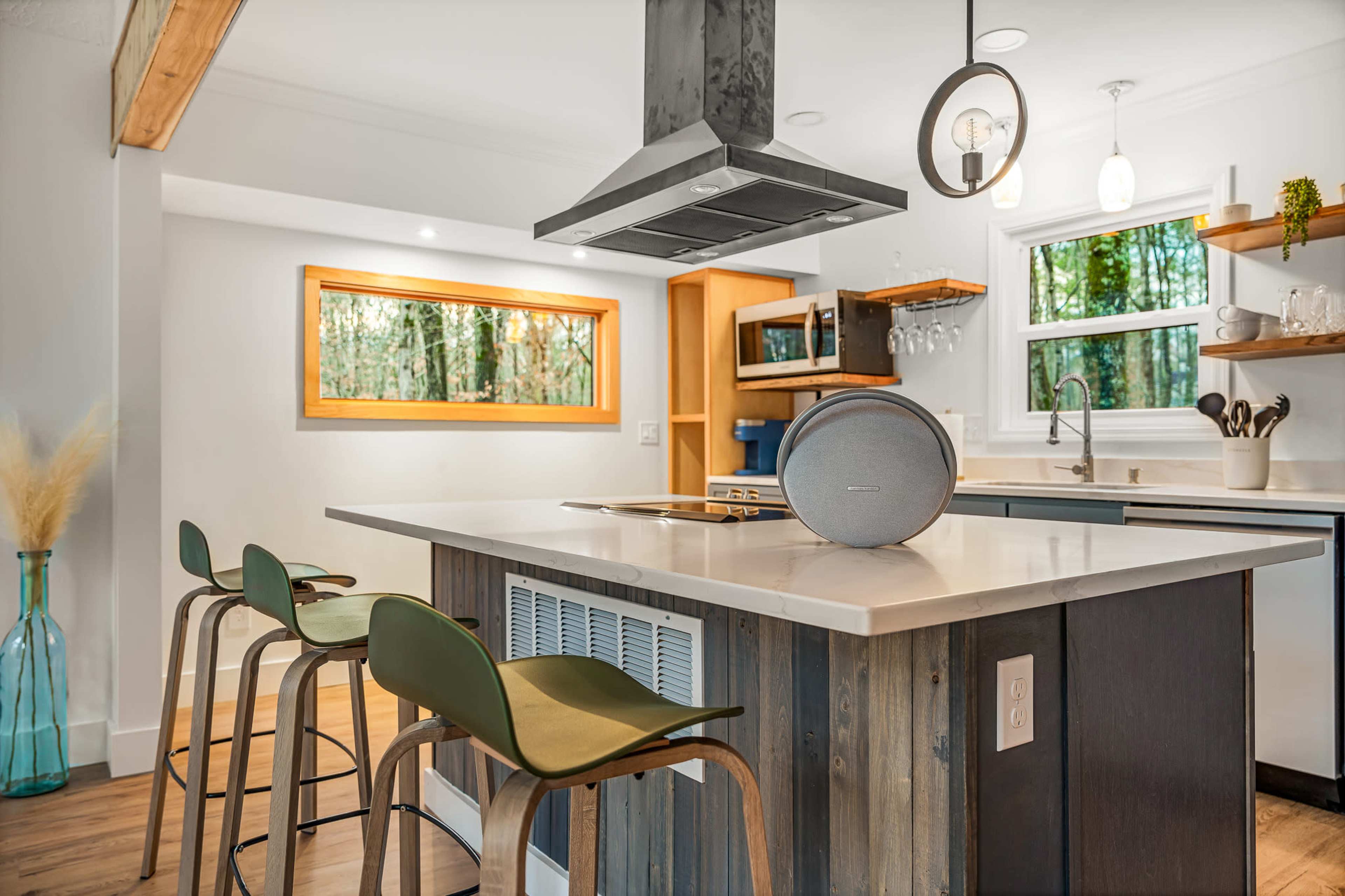 A modern kitchen features a central island with a stovetop and sleek bar stools, illuminated by natural light streaming through a window overlooking a wooded area.
