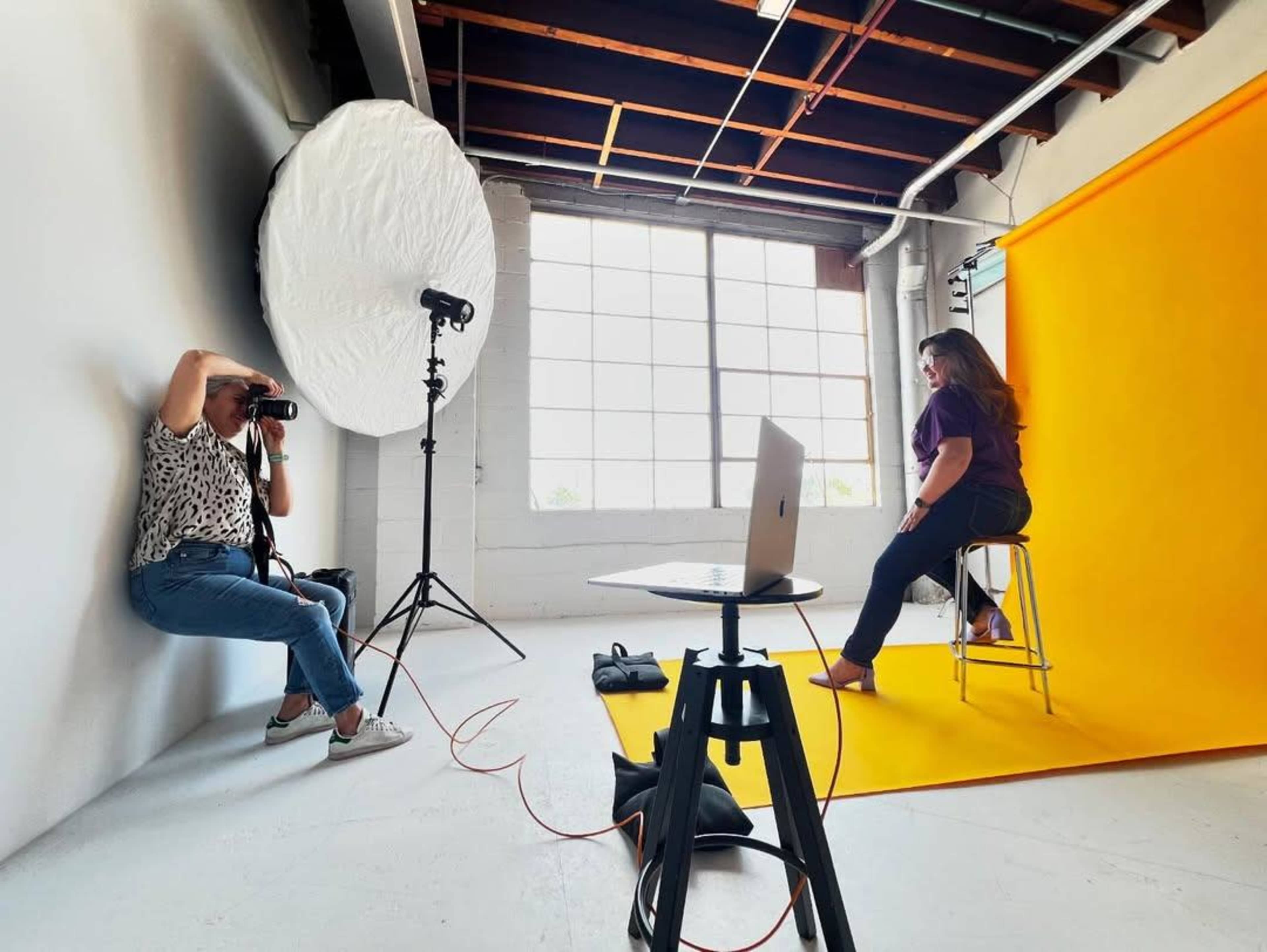 A photographer is capturing images of a model sitting on a stool against a bright yellow backdrop in a well-lit studio.