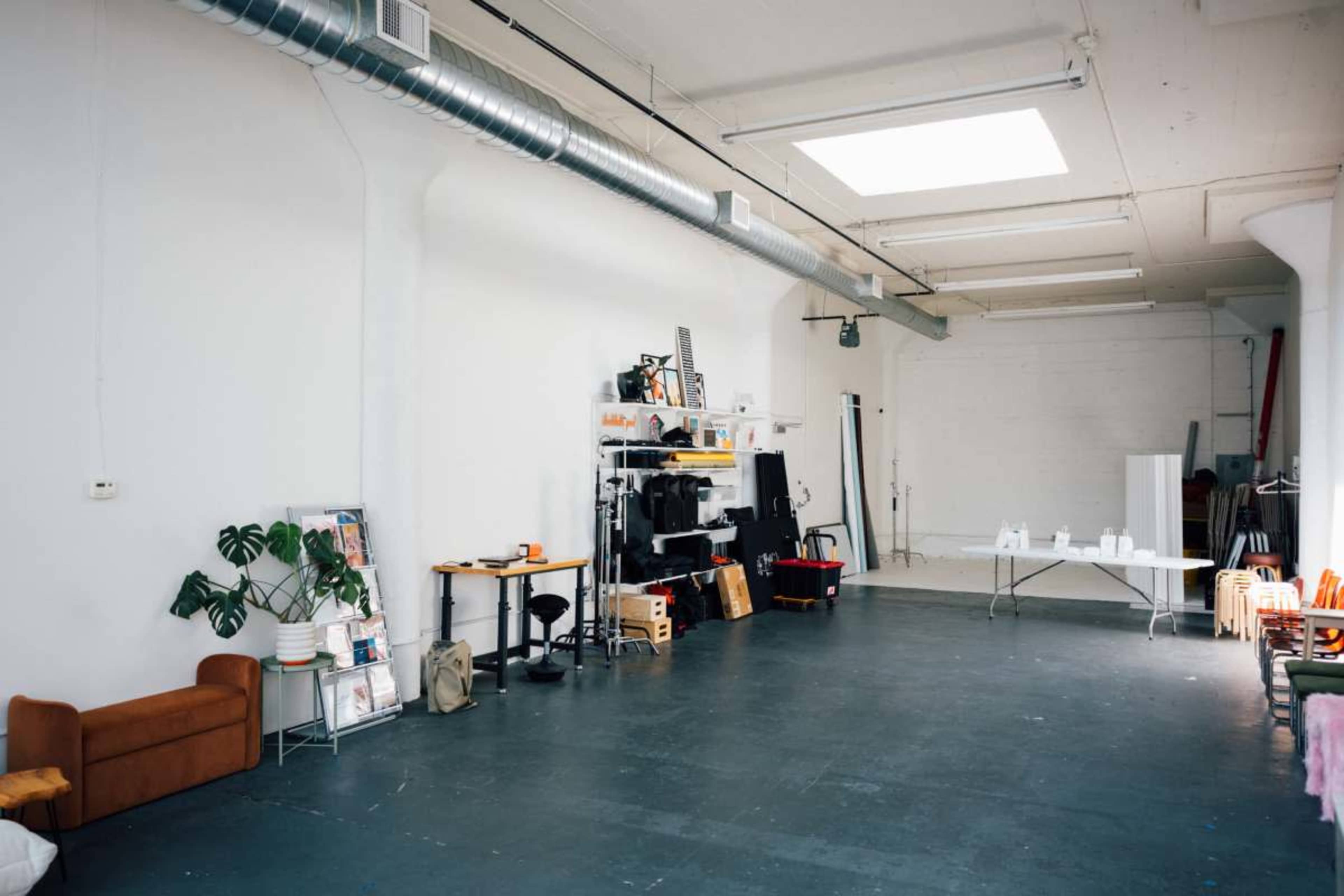 The image shows a spacious, empty studio with a grey floor, a couch, shelving units, and a table, illuminated by natural light from a skylight.