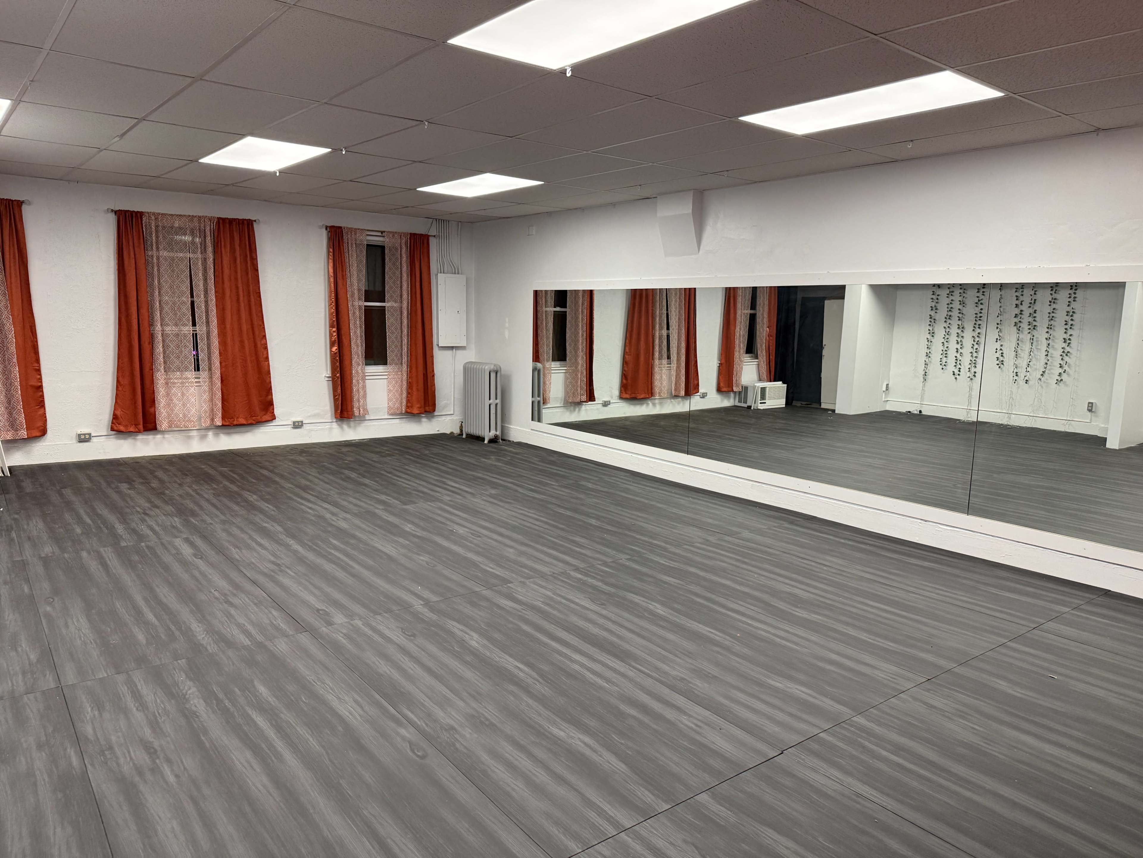 The image shows an empty dance studio with gray flooring, white walls, and large mirrors reflecting orange curtains at the windows.