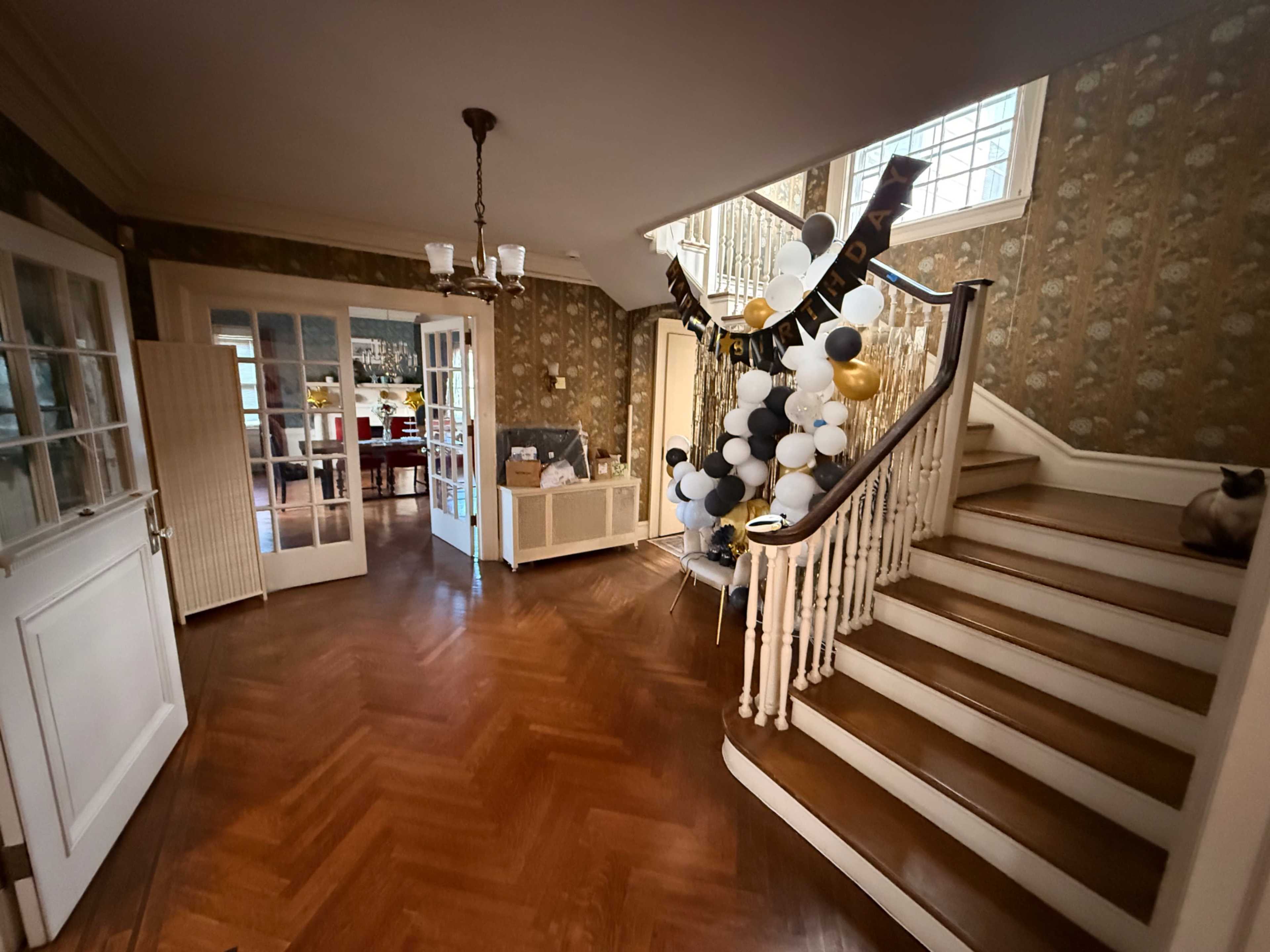 The image shows a spacious foyer with a wooden staircase, adorned with a decoration of black, white, and gold balloons, and features an adjacent room visible through double doors.