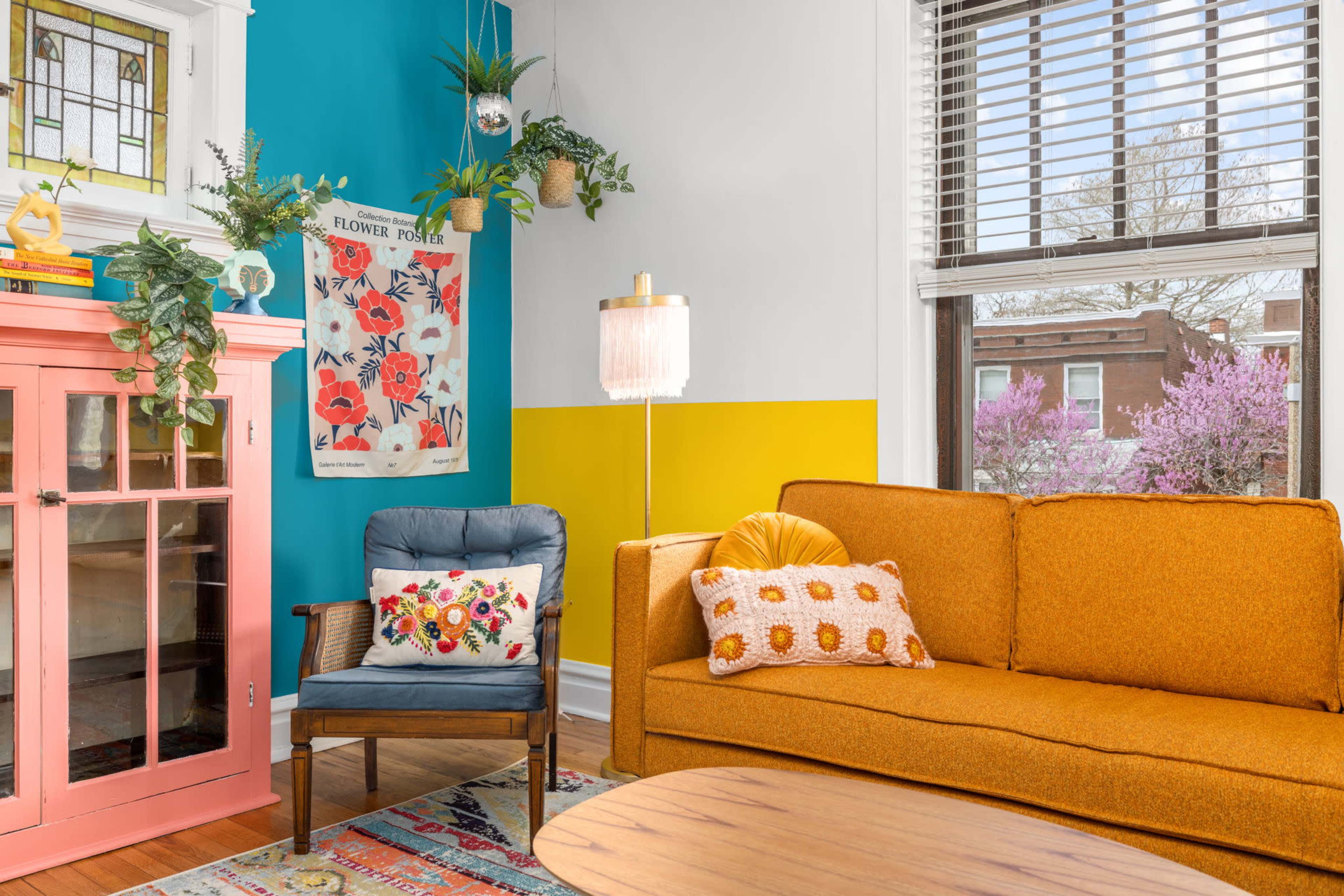 The scene features a brightly decorated living room with an orange sofa, a blue armchair, and colorful wall art, complemented by plants and a large window.