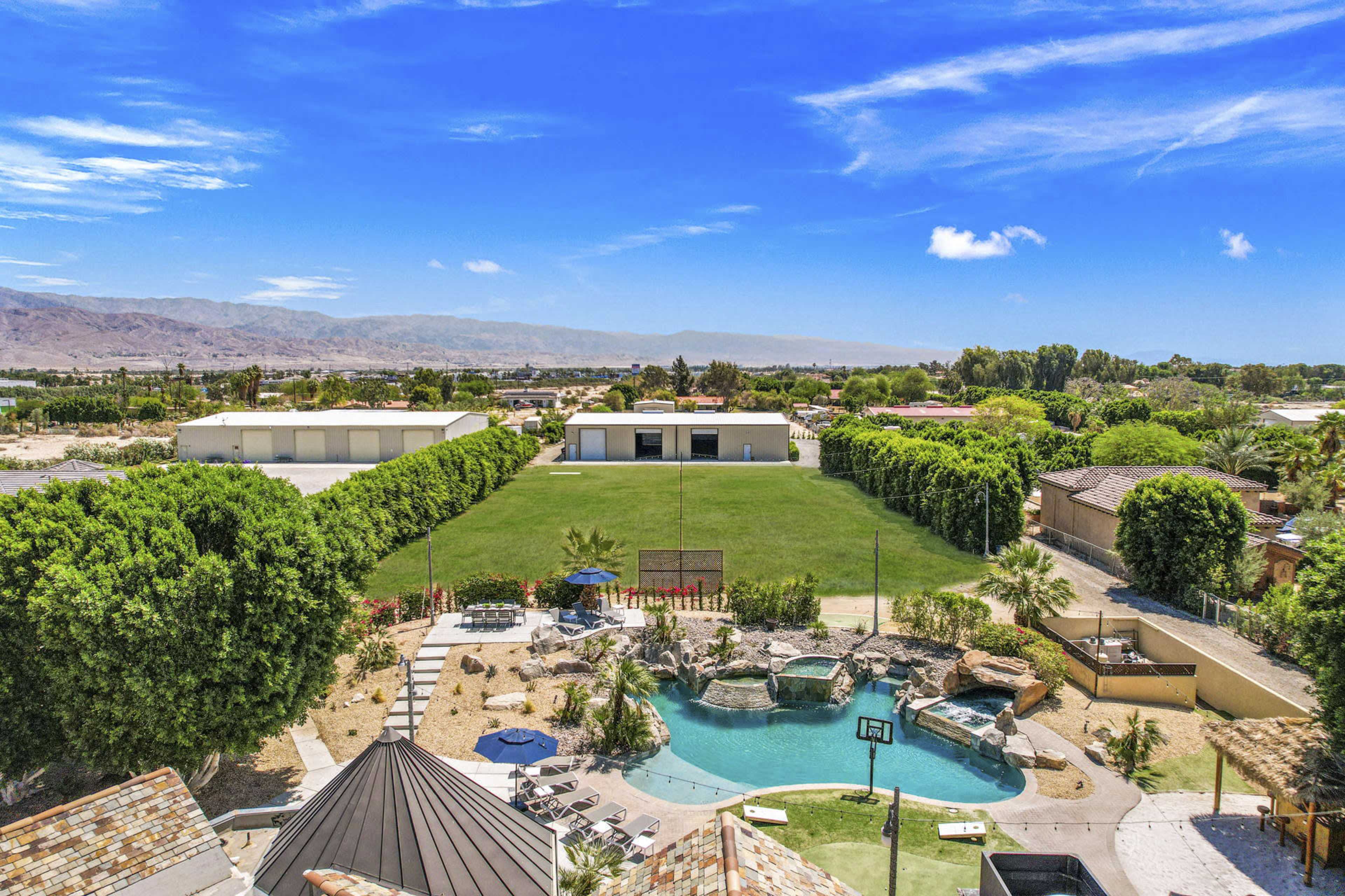 A spacious outdoor area featuring a large green lawn, several palm trees, a swimming pool with a rock formation, and multiple buildings in the background against a clear blue sky.