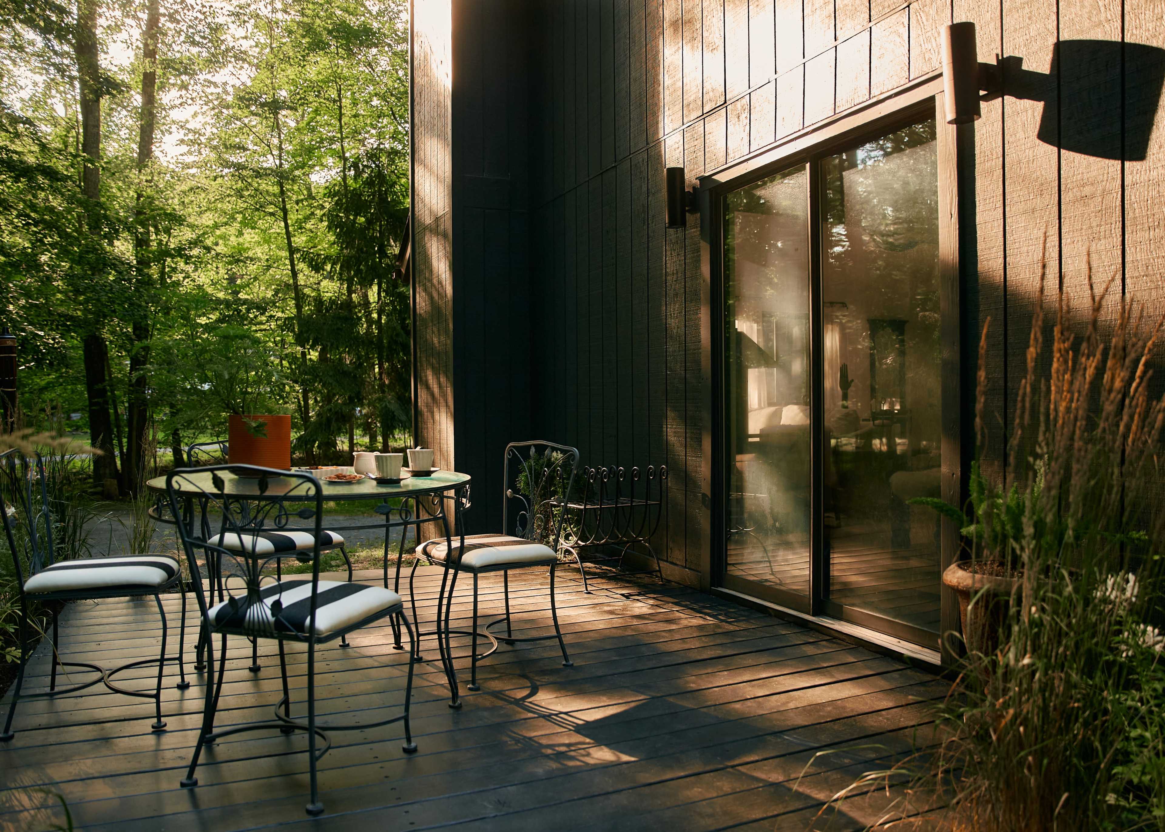 A set of metal chairs and a round table are arranged on a wooden deck beside a glass door, surrounded by greenery in soft afternoon light.