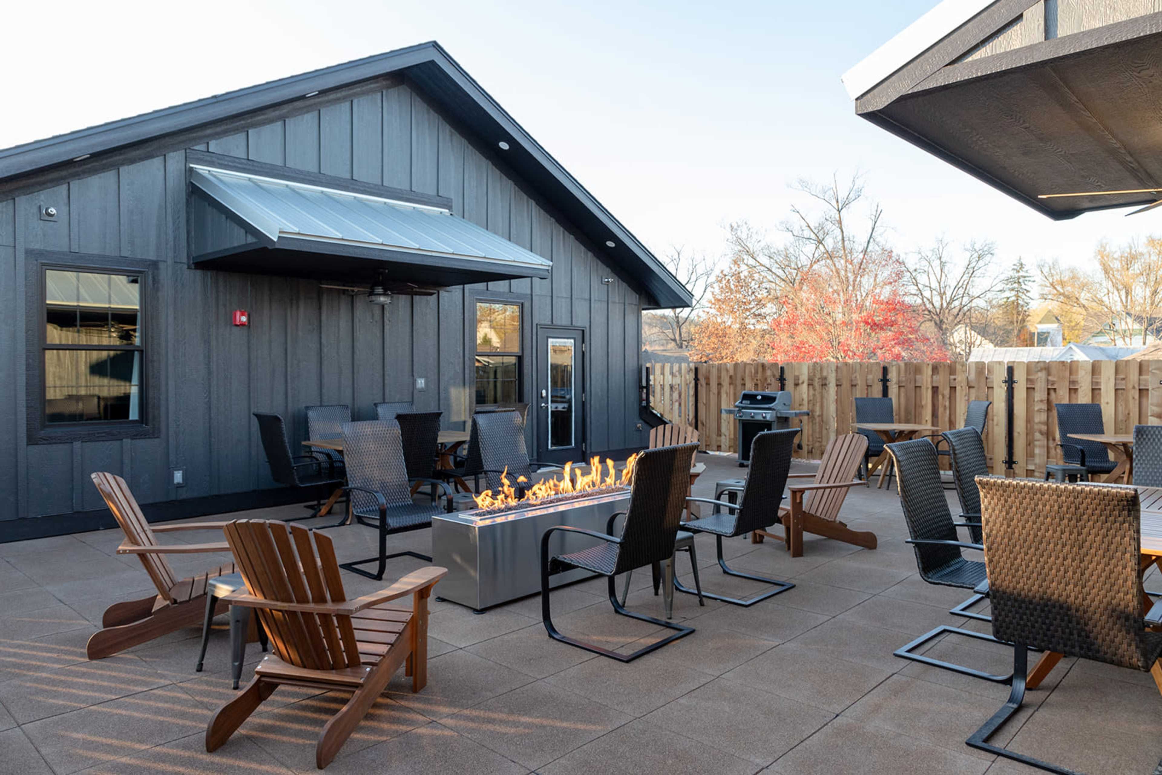The image shows a patio area with several outdoor chairs arranged around a central fire pit, adjacent to a wooden fence and a building in the background.