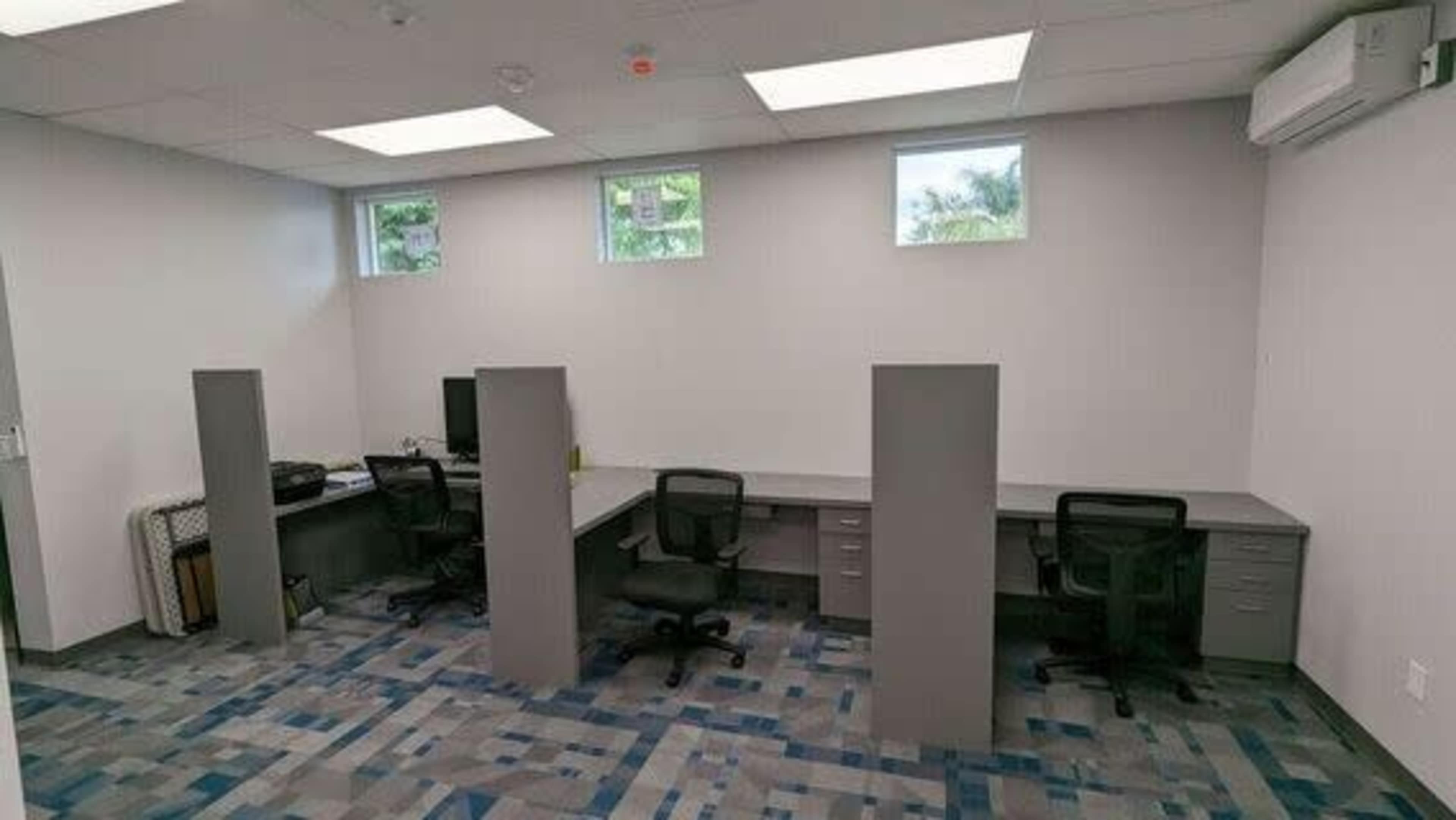 A modern office space with four desks separated by gray dividers, two chairs per desk, and a computer setup in one corner.