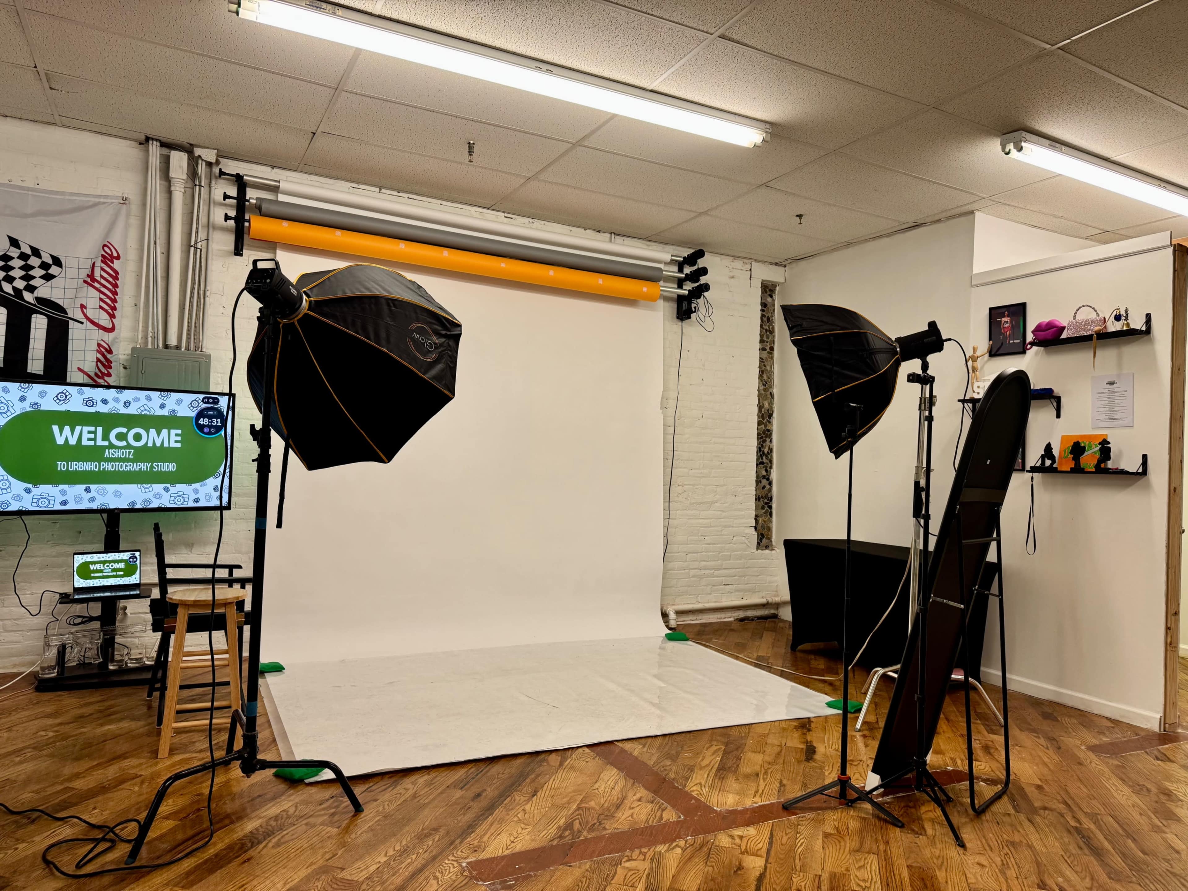 The image shows a photography studio with a white backdrop, two large light umbrellas, a monitor displaying a welcome message, and various props and a black table on the side.