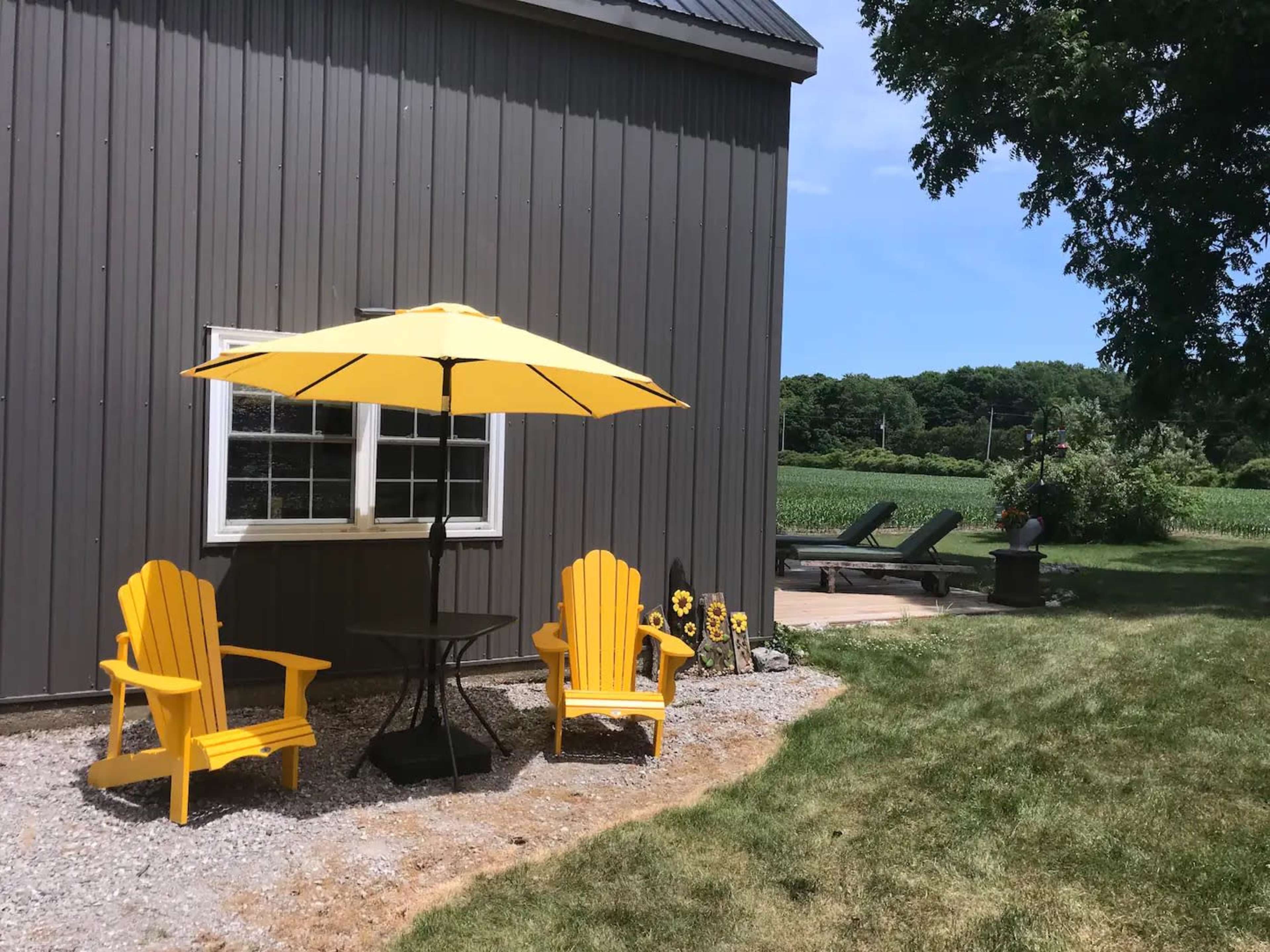 Two yellow chairs and a table sit under a yellow umbrella next to a gray building, with a grassy area and a view of a field in the background.