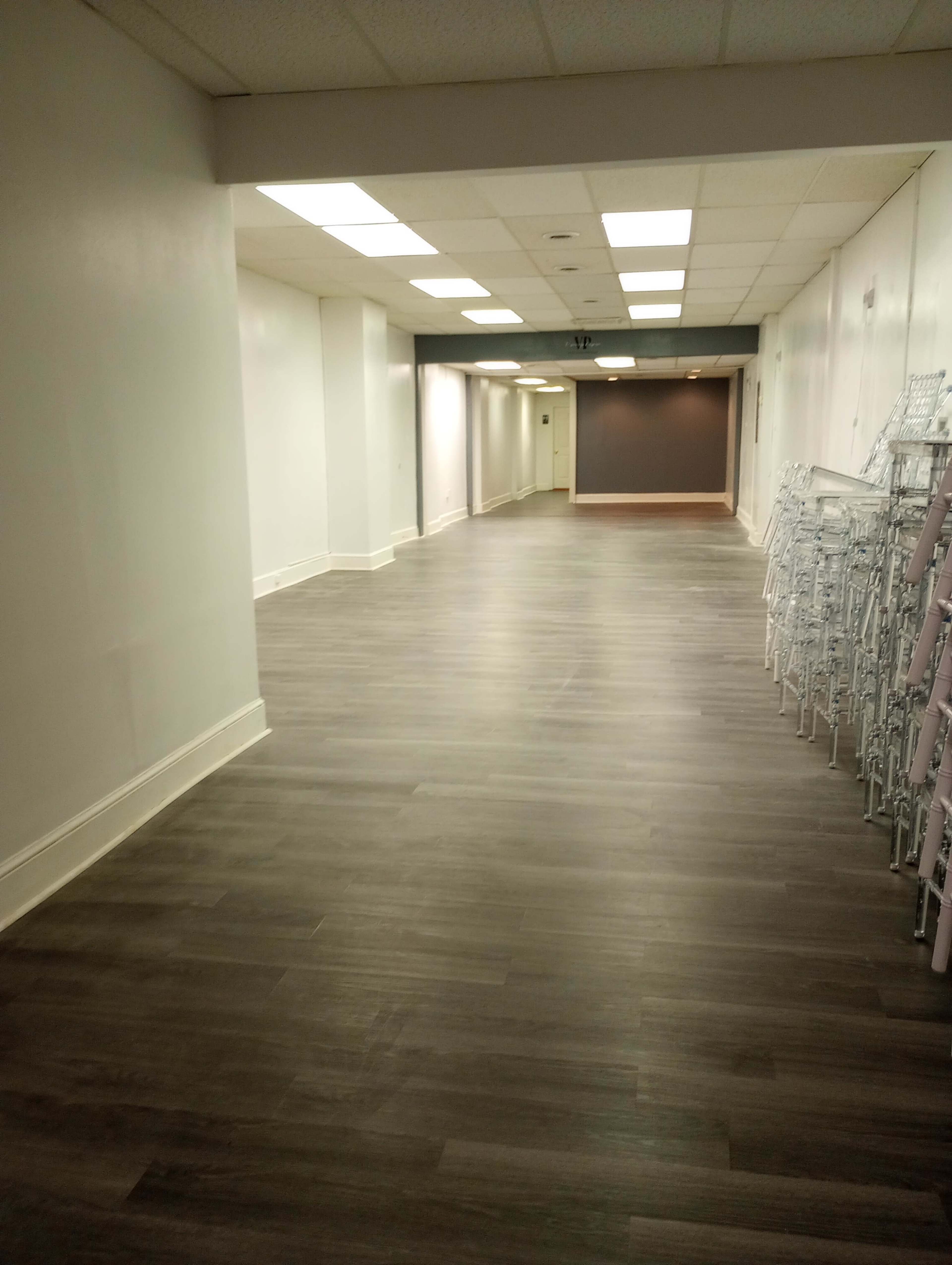 A long, empty corridor with wooden flooring and a row of folding metal chairs on the right side.