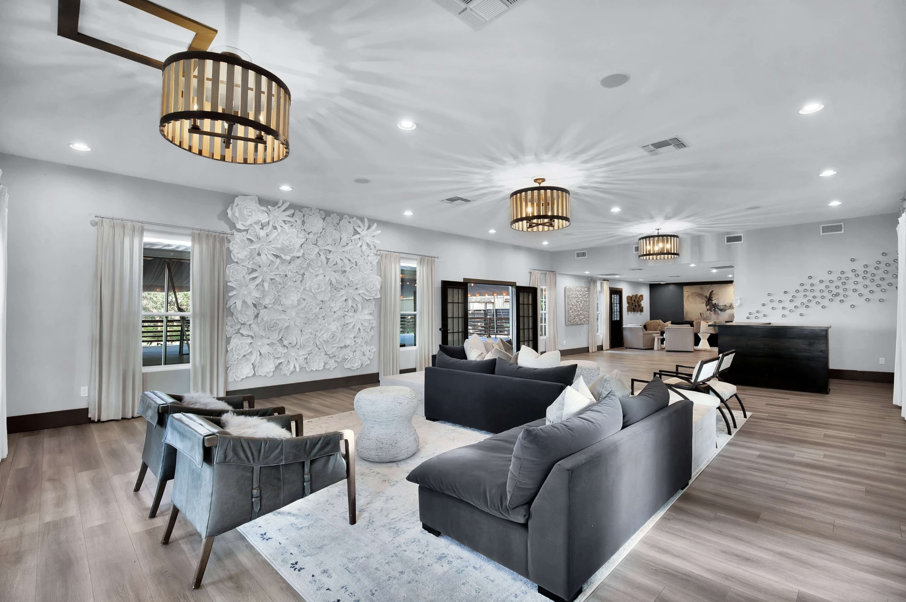 A modern living room with two dark sofas, several decorative chairs, a light rug, and large windows, complemented by unique light fixtures and wall decorations.