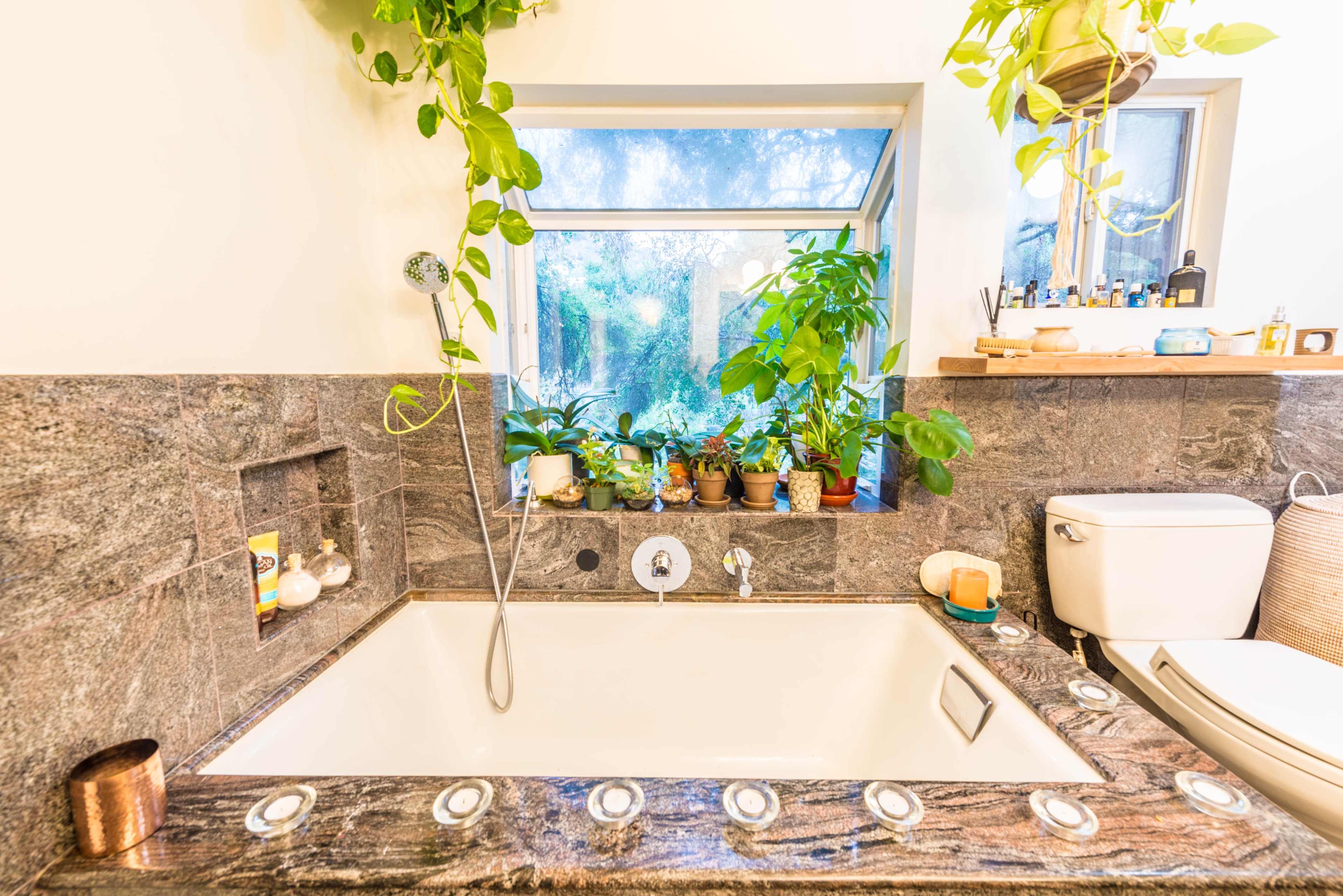 A modern bathroom featuring a stone bathtub surrounded by plants and natural light streaming through a large window.