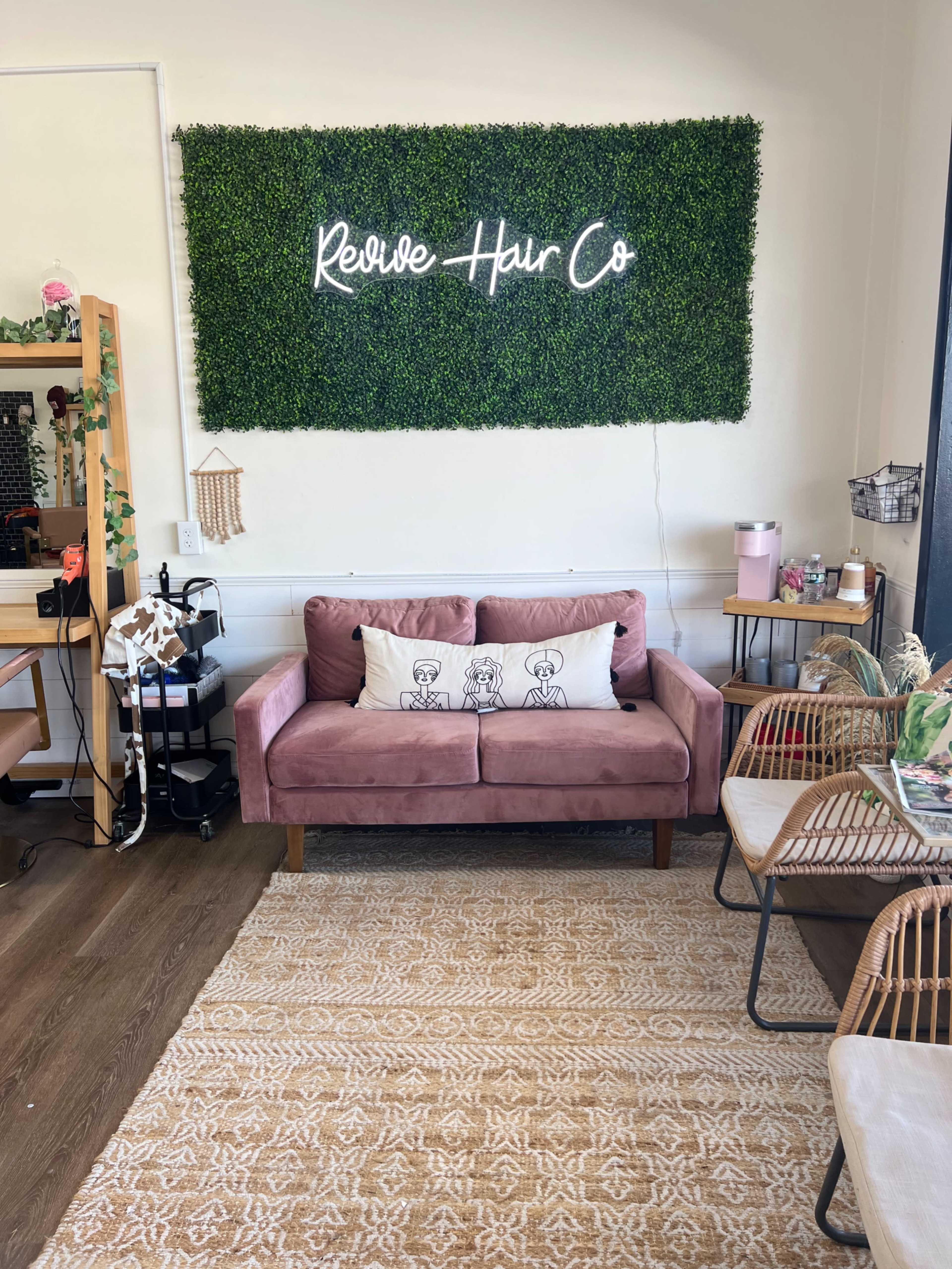 A cozy salon space featuring a pink couch with decorative pillows, a green wall with a "Revive Hair Co" sign, and several stylish seating areas.
