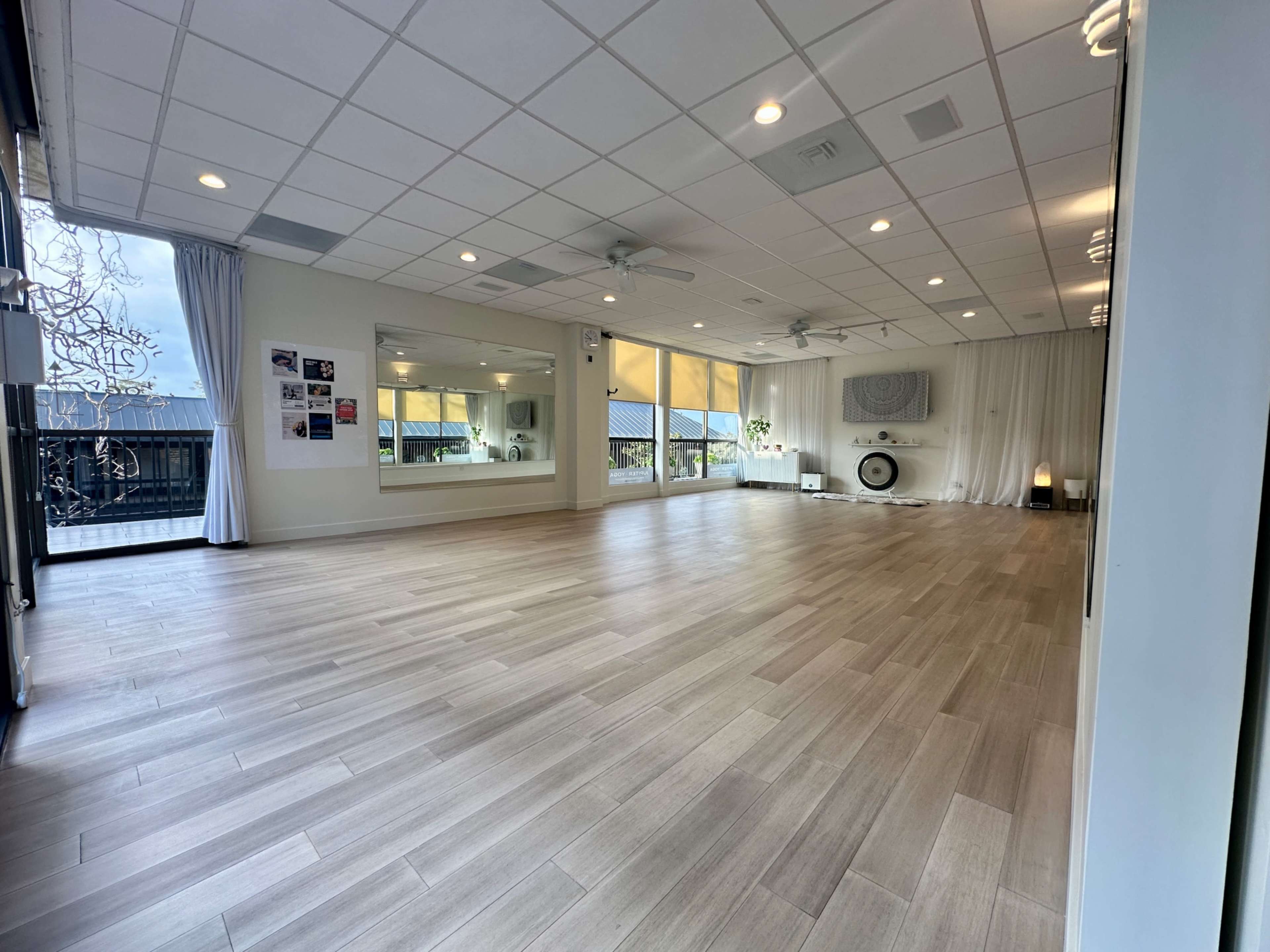 The image shows a spacious, well-lit interior of a yoga or fitness studio with wooden flooring and large windows.