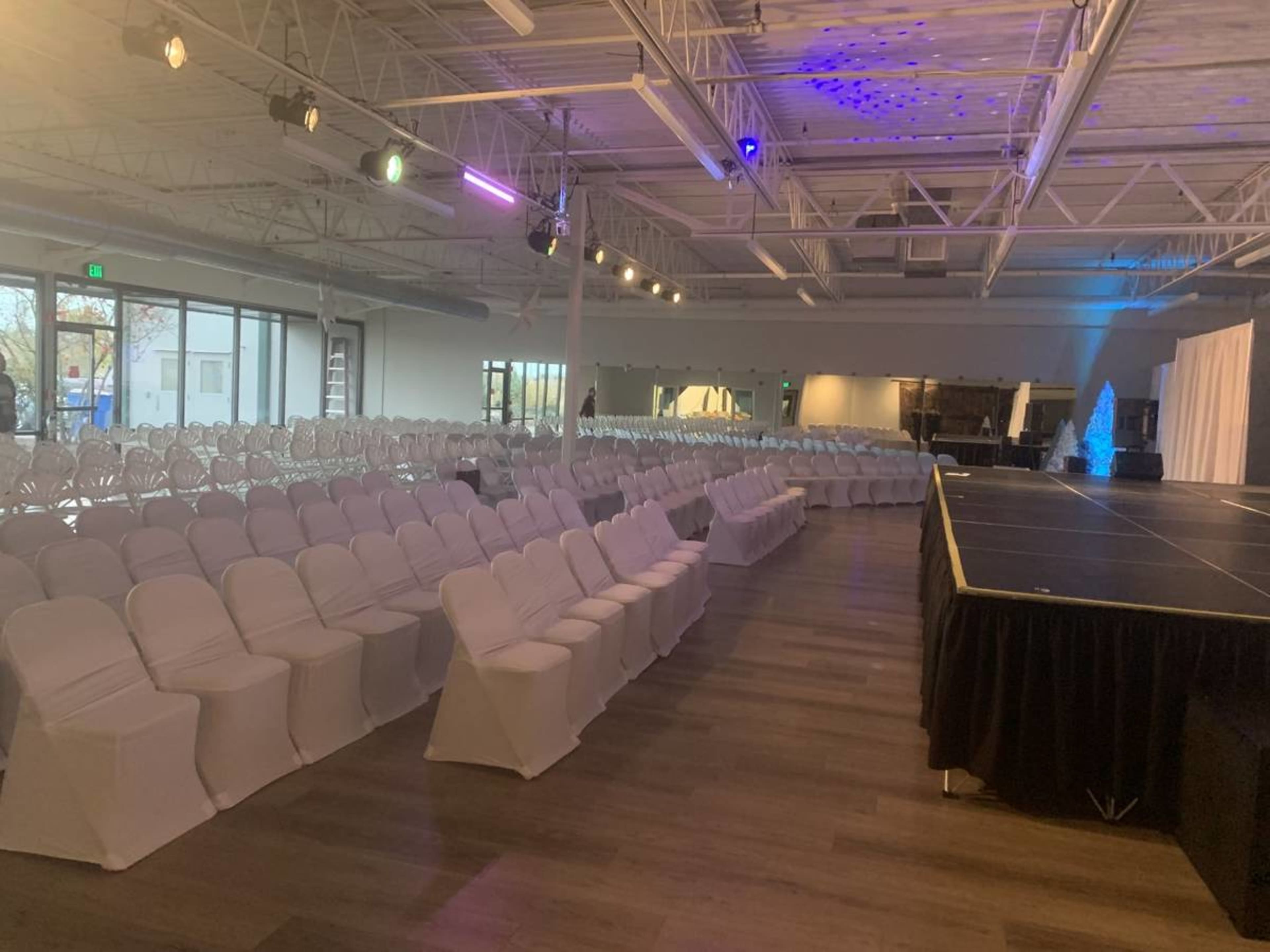 A spacious event venue is set up with rows of white chairs facing a black stage, illuminated by lights from the ceiling.