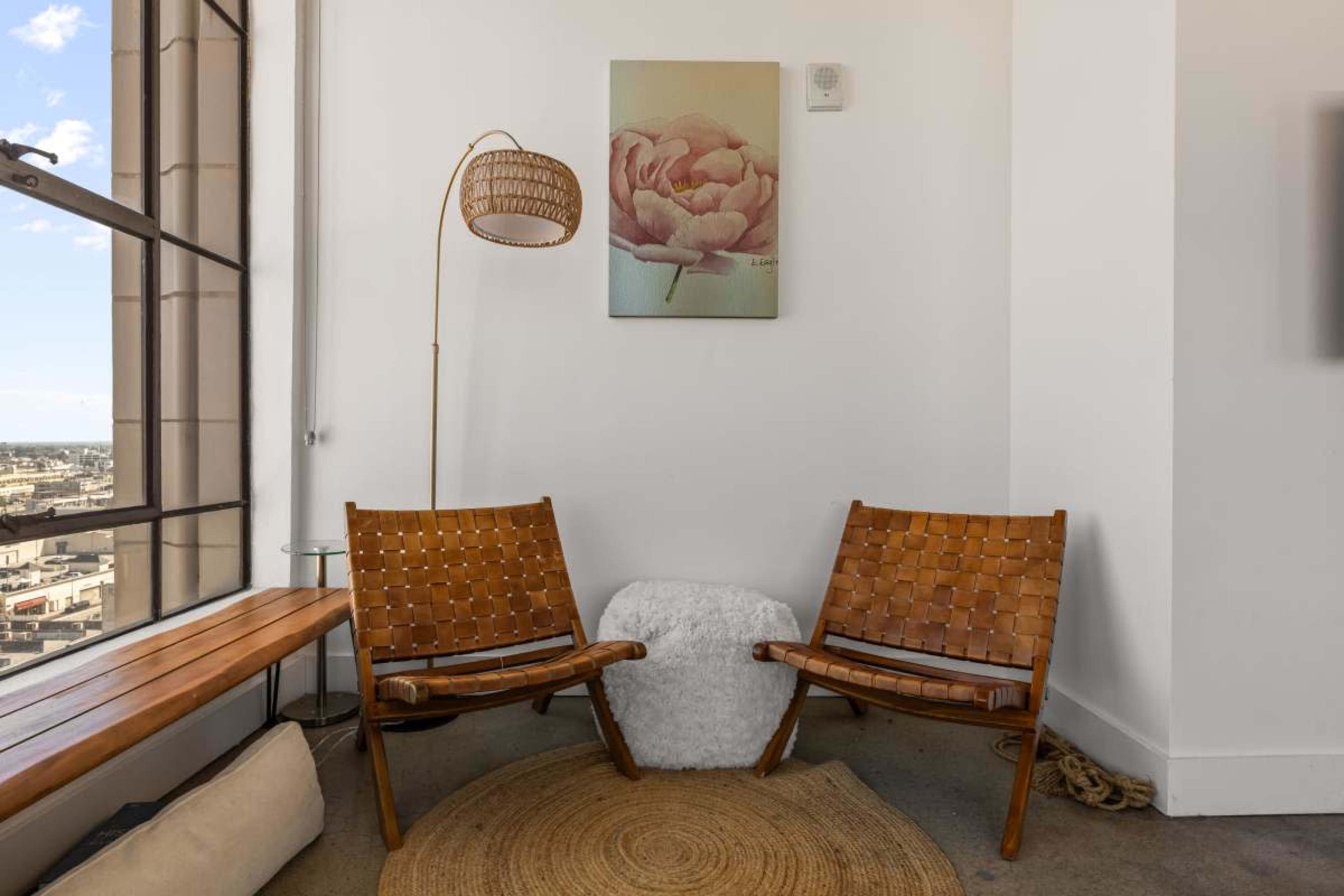 A corner of a room features two woven wooden chairs, a round white side table, a lamp, and a peony painting on the wall.
