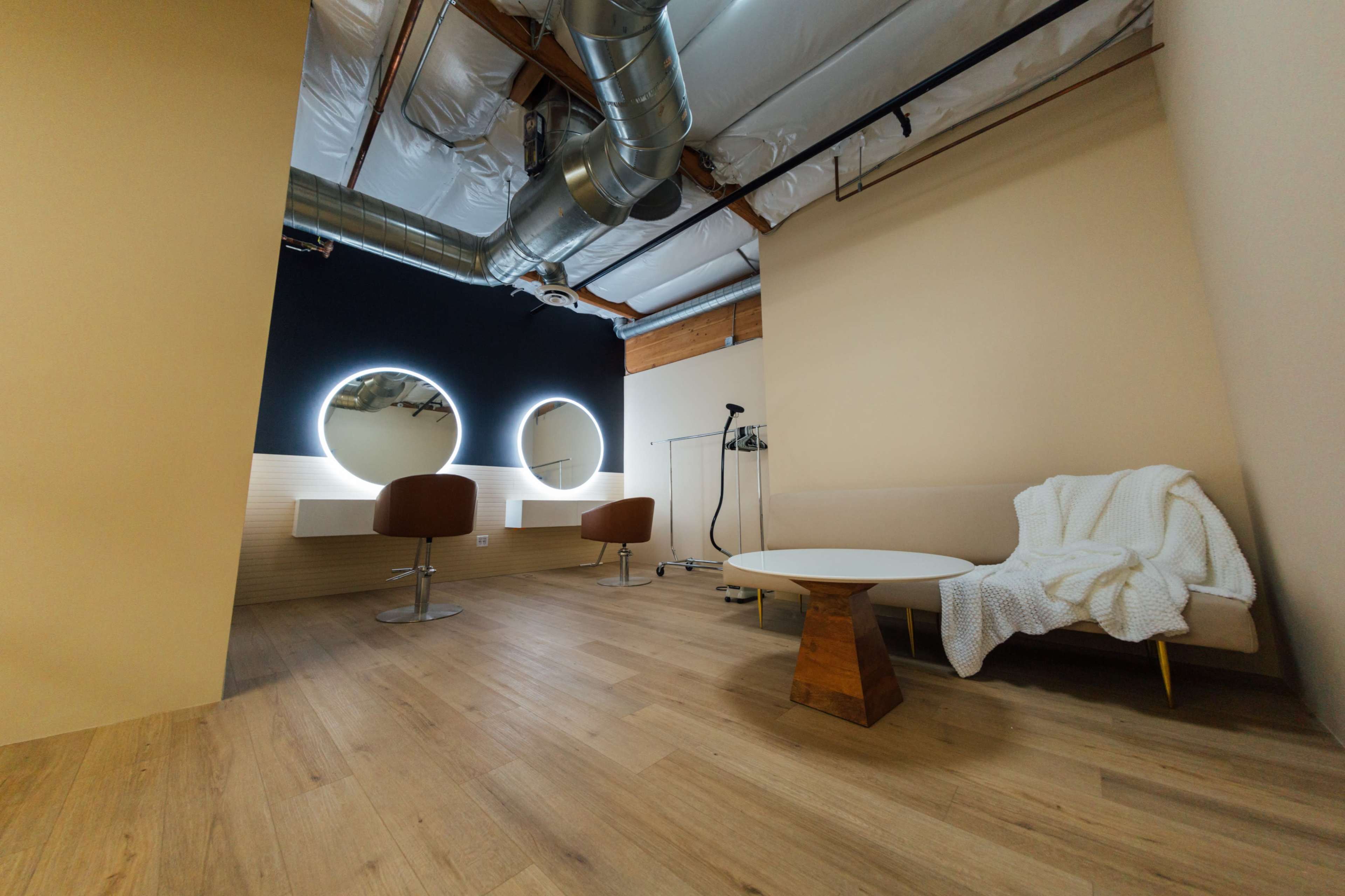 The image shows a minimalist room with two circular mirrors, a beige couch, a wooden coffee table, and exposed ductwork on the ceiling.