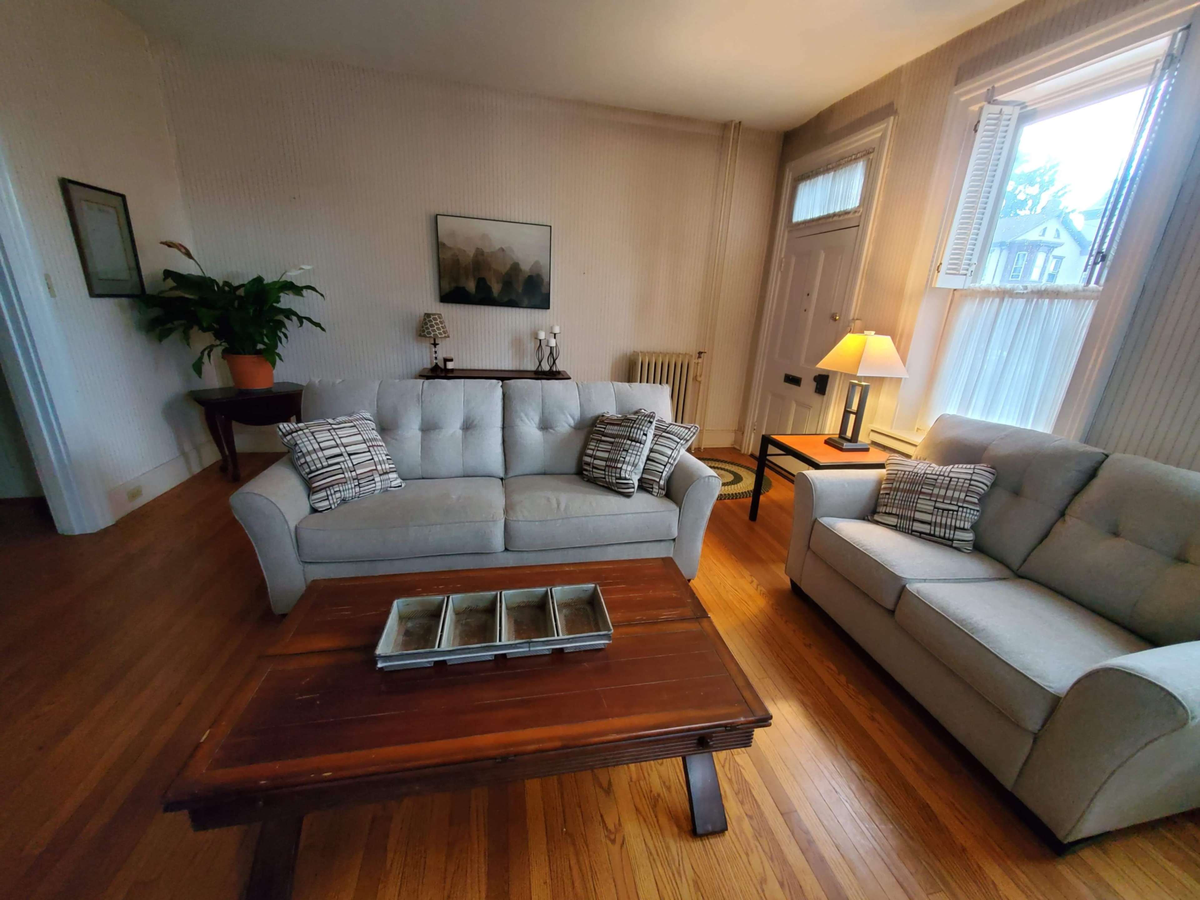 The living room features a light-colored sofa and loveseat arranged around a wooden coffee table, with a potted plant in the corner and a lamp on a side table.