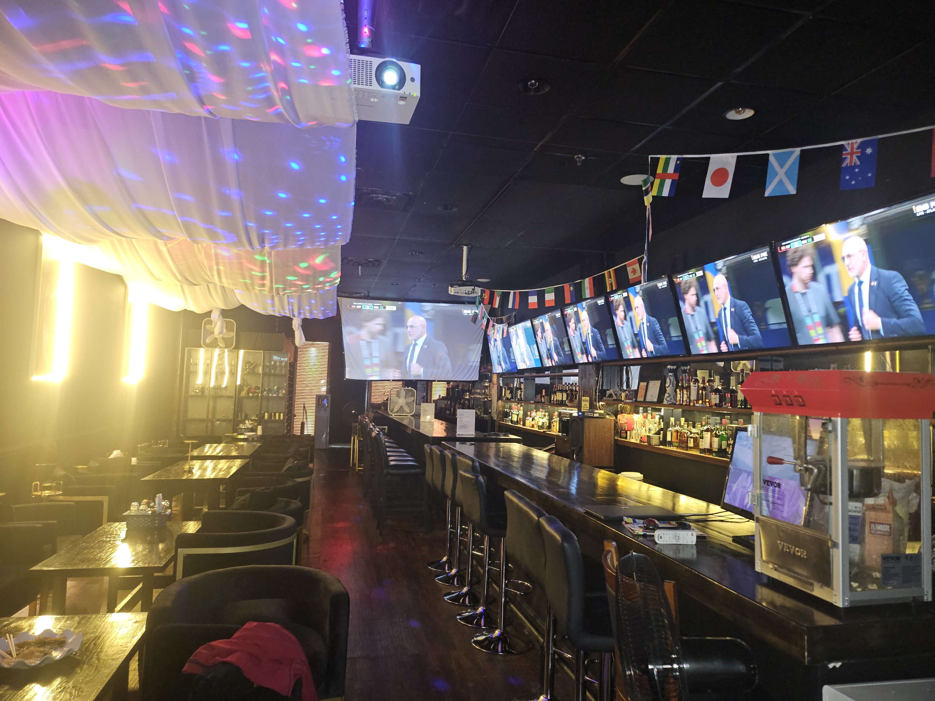 The image shows a dimly lit bar with a long counter, multiple televisions displaying a sports event, and colorful lights projected on the ceiling.