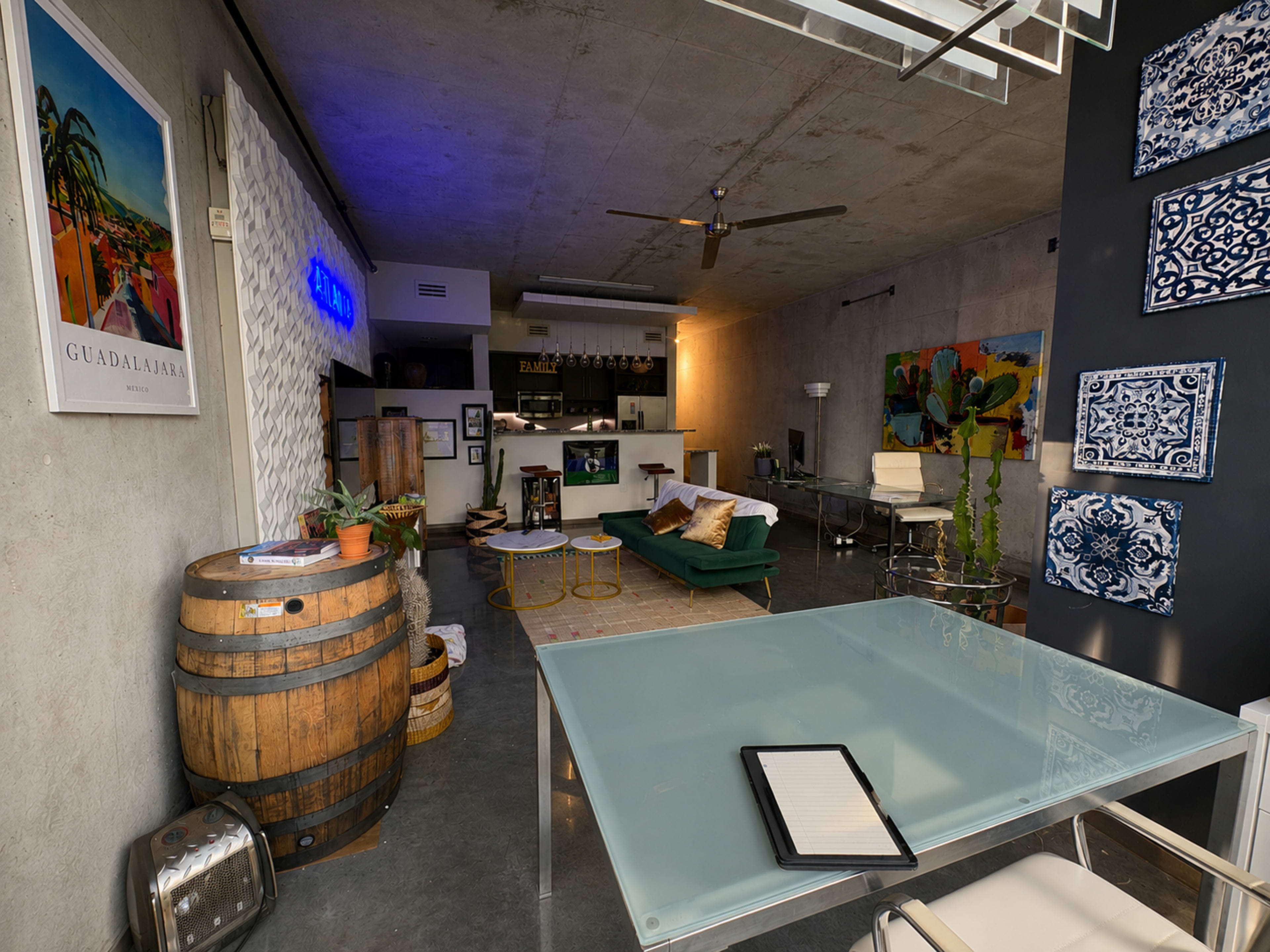 The image shows a modern, industrial-style living space featuring a green couch, a glass dining table, and a wooden barrel, with artwork and a bar area in the background.