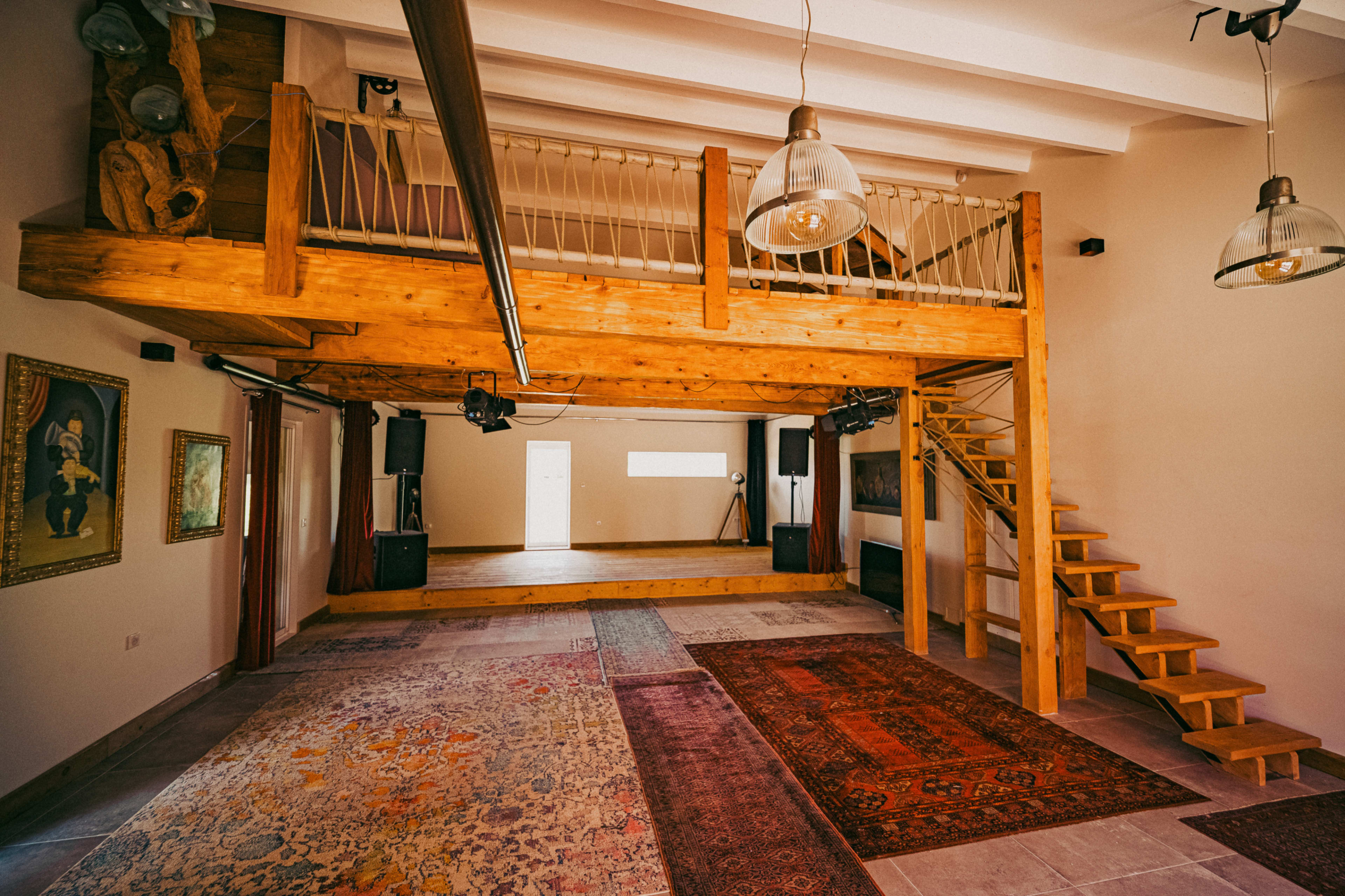 The image shows a spacious, well-lit room featuring a wooden loft, a staircase, and several rugs on the floor.