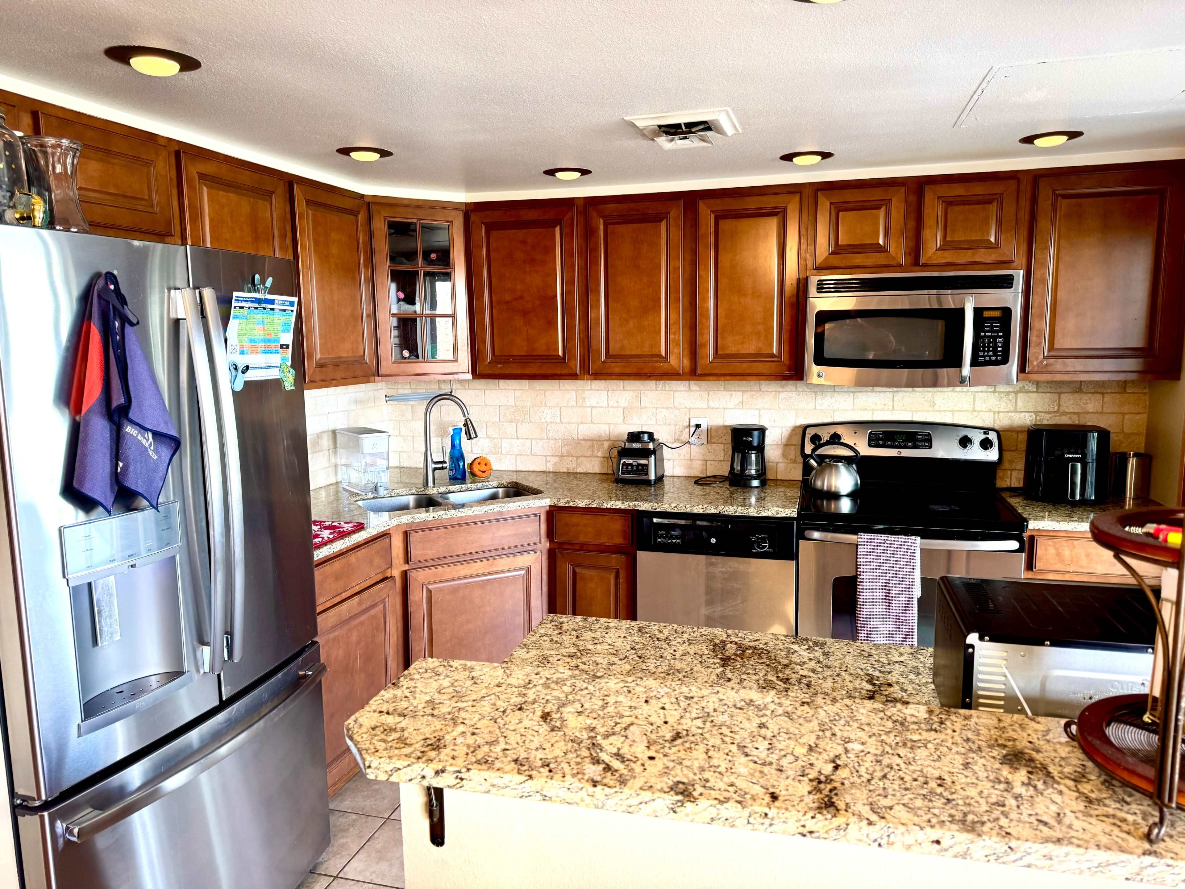 The image shows a modern kitchen with wooden cabinetry, stainless steel appliances, and a granite countertop.