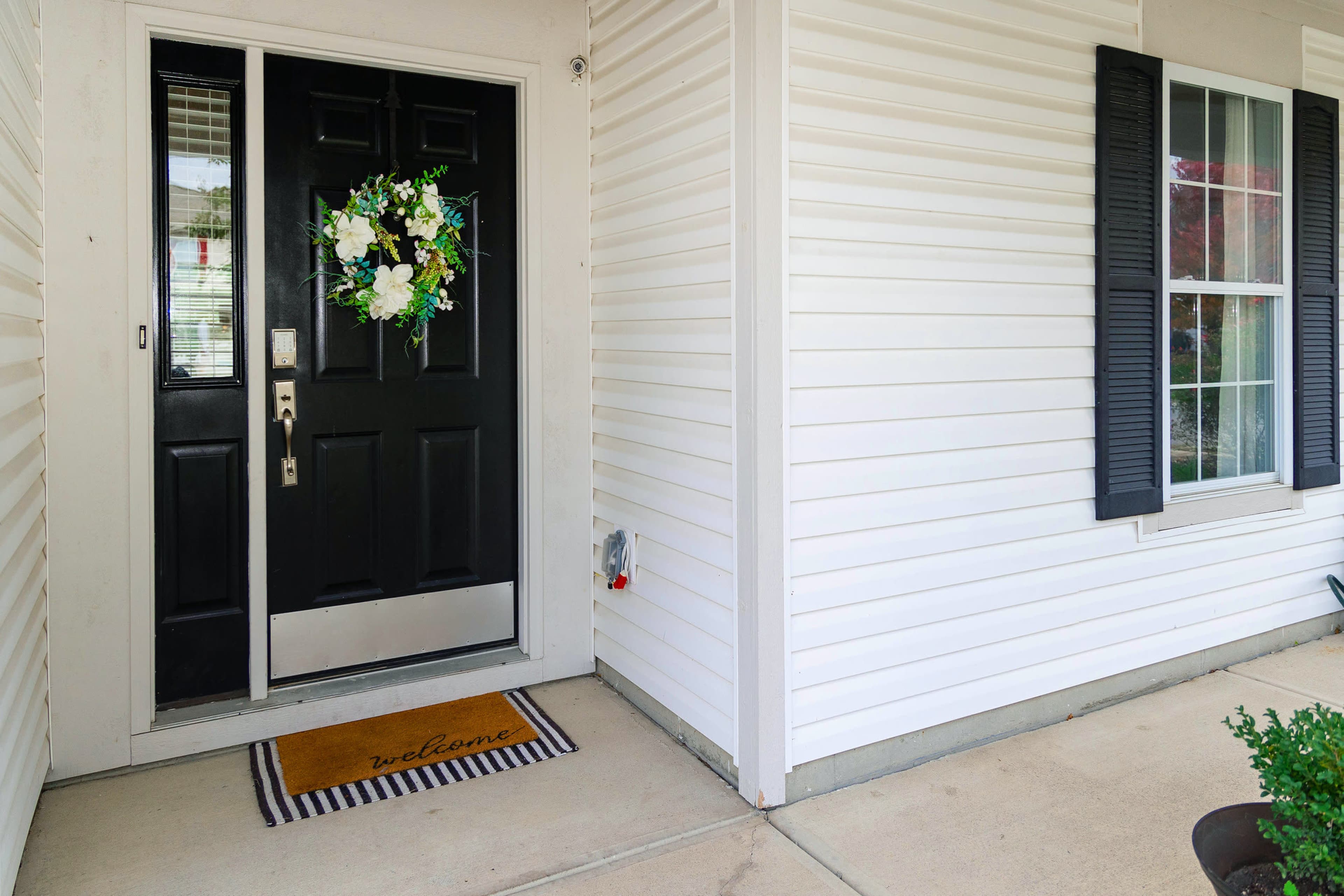 A black front door adorned with a floral wreath, with a "welcome" doormat placed on a concrete porch beside a house with white siding and black shutters.