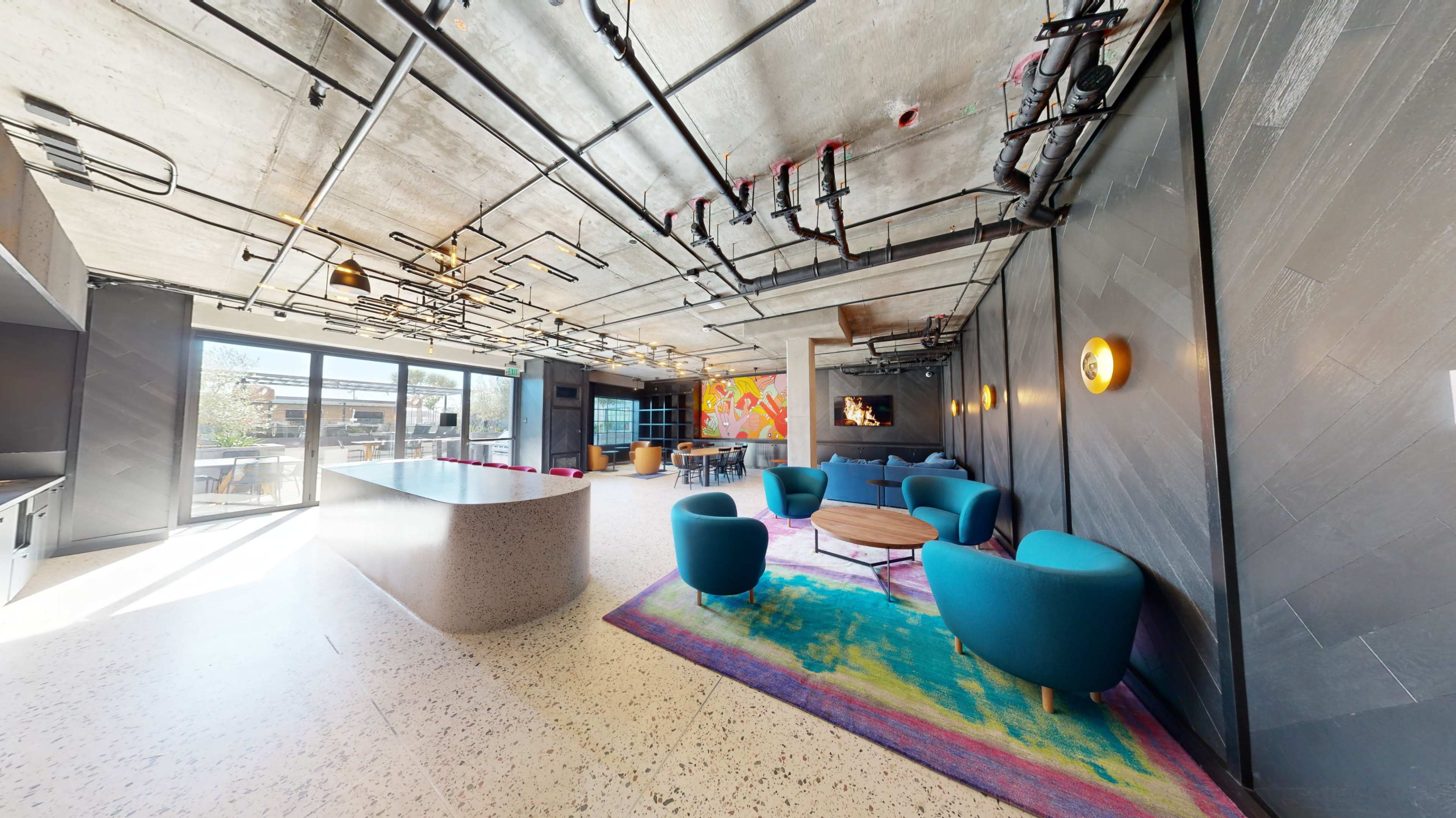 The interior space features an open layout with modern furniture, including colorful chairs and a central round table, alongside large windows that provide natural light.